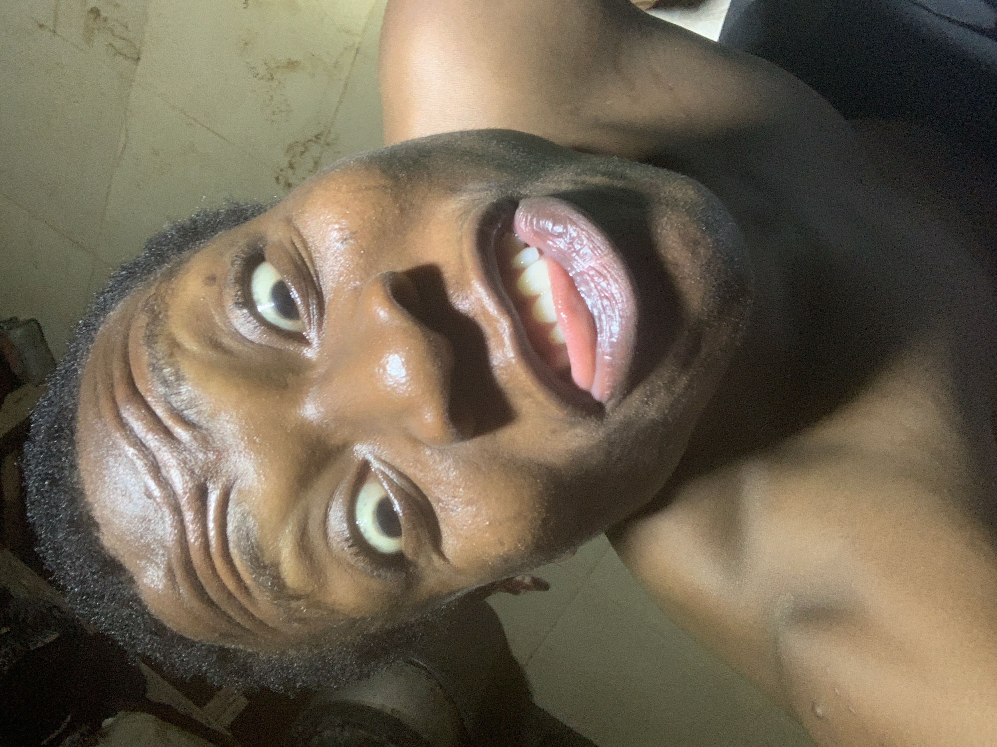 A close-up, low-angle shot of a shirtless dark-skinned man with wide eyes, dark curly hair, and a slightly open mouth, revealing his teeth and tongue. The background appears to be a lightly colored wall or floor. The man is oriented sideways, with his face tilted toward the viewer. The location is Unknown, Unknown.