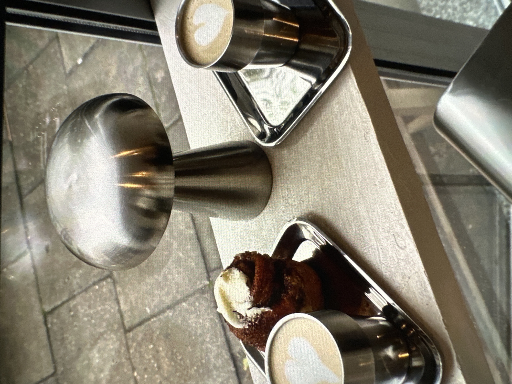 An overhead shot depicts two servings of coffee, each on a square metal tray, accompanied by a pastry. The scene is viewed from a location in Yerevan, Armenia. A metal object with a spherical end sits near one of the trays. The coffee in each cup appears to have latte art. The pastry, partially covered in a white glaze, rests on one of the trays next to a coffee cup. The photograph is taken on or near a surface adjacent to a window.