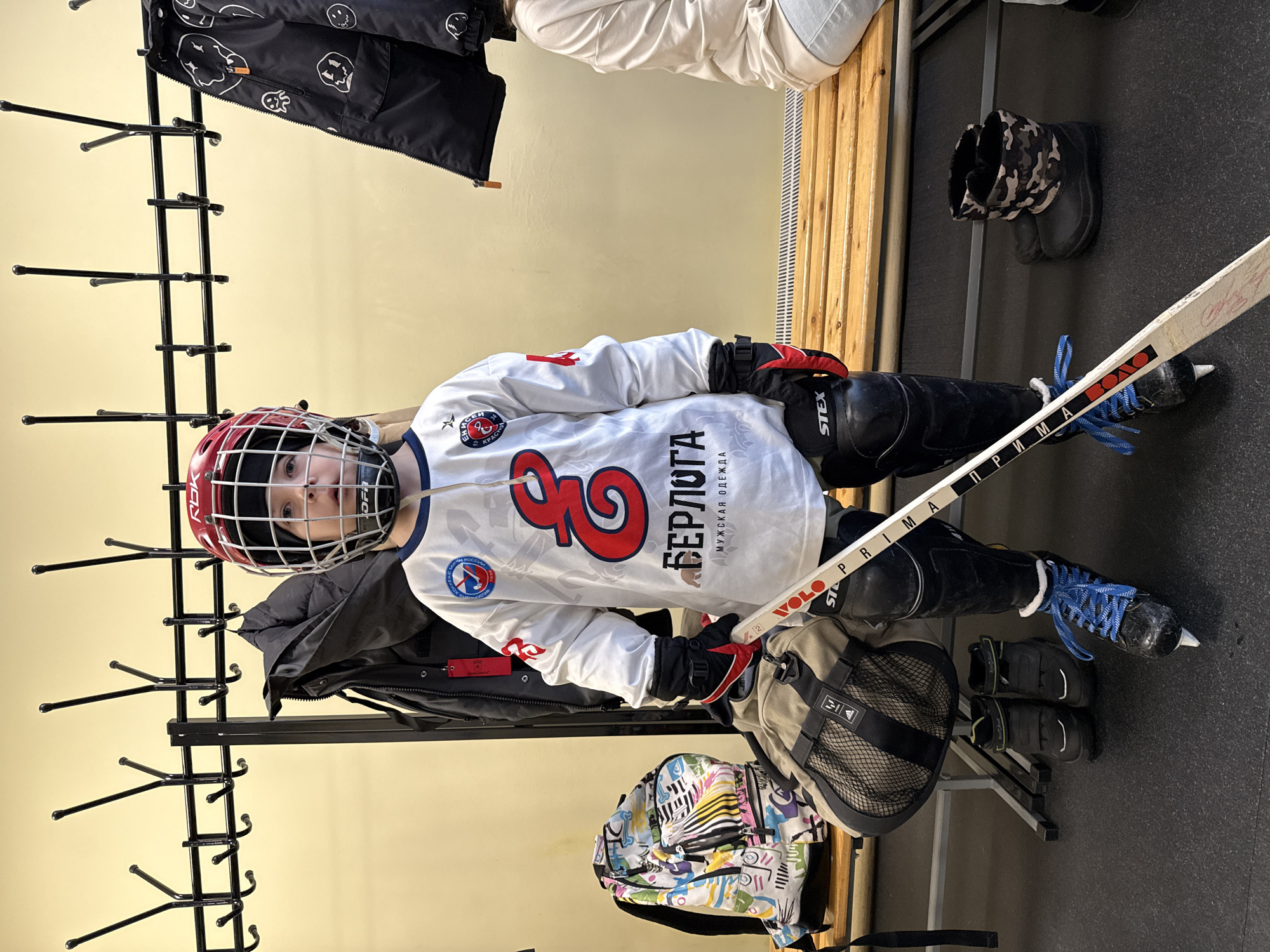 A child in full hockey gear, including a red helmet with a face cage, white jersey with number 3 and the word "БЕРЛОГА" (BERLOGA) on it, knee pads, gloves, and blue-laced skates, sits on a wooden bench in what appears to be a locker room. A hockey stick rests beside them. Various clothing items hang on a black metal coat rack to the left. A backpack with colorful designs sits on the floor, and another bag is near the bench. Boots are visible on the floor to the right of the bench. The jersey displays a patch of the Krasnoyarsk hockey team, so the location is most likely in Krasnoyarsk, Russia.