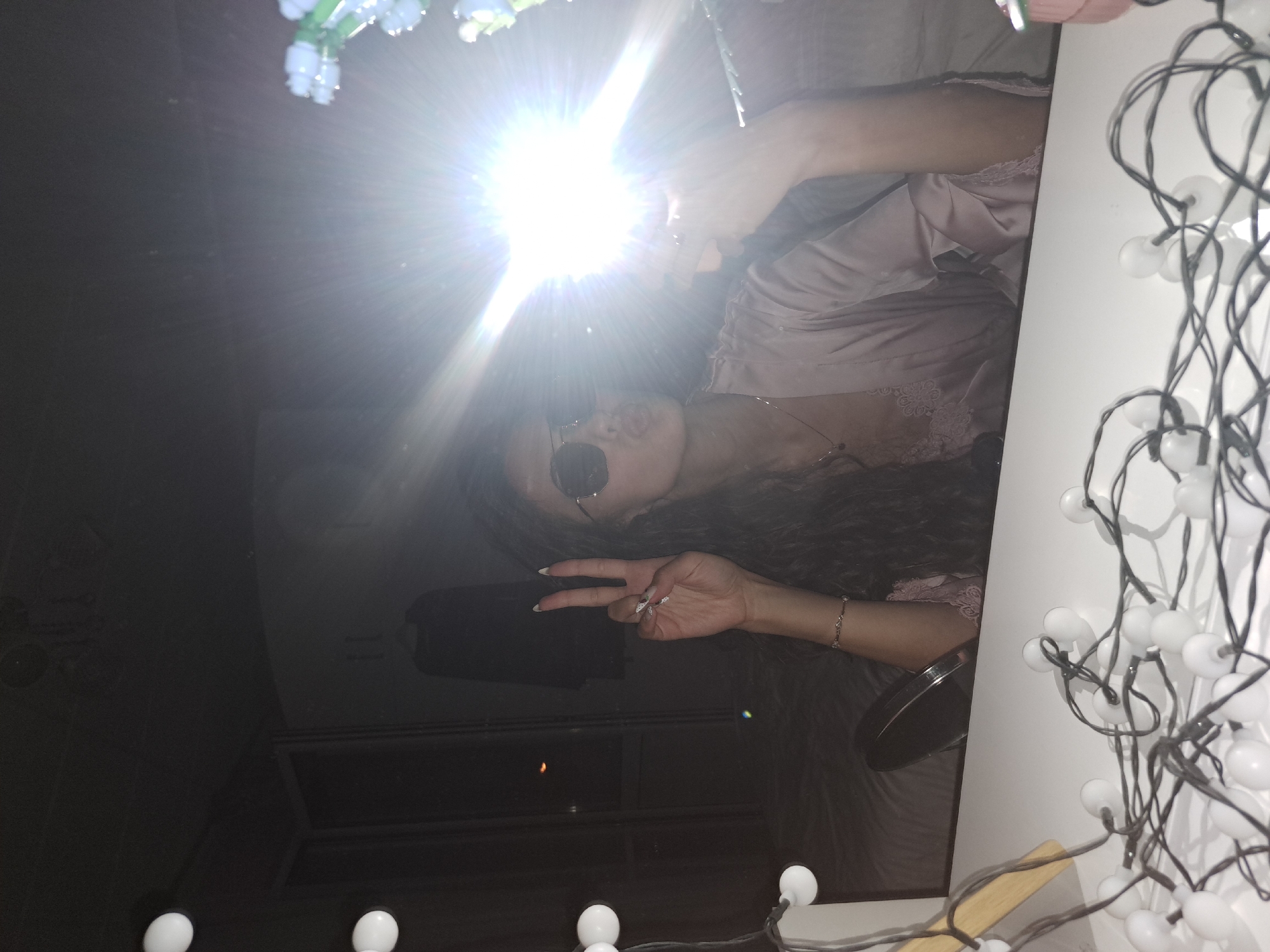 The image captures a playful mirror selfie, likely taken in a dimly lit indoor setting such as a bedroom in Negovan, Bulgaria. The primary subject is a person, likely female, with long, dark, curly hair, seen in reflection. She is wearing sunglasses and a light-colored, possibly pink or lavender, satin robe adorned with lace trim.
The person is holding a smartphone, whose flash creates a prominent, bright starburst effect that illuminates her upper body and face. Her lips appear slightly puckered, suggesting a kissy face. Her right hand is raised, making a "peace" sign (V-sign), and her fingernails are manicured with a light polish, possibly with a subtle design. A delicate bracelet is visible on her wrist.
To the left of the subject, a string of white, spherical fairy lights with dark wires is draped, resting on a light-colored surface, possibly a vanity or shelf. The background reflected behind the person is dark and indistinct, revealing faint architectural elements that suggest the interior of a room, possibly at night or in very low light. The image conveys a casual and confident mood, capturing a self-portrait moment indoors. There is no visible text in the image.