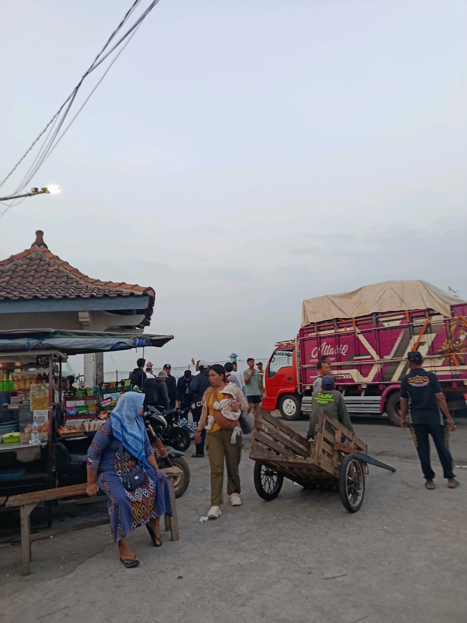 The image depicts a street scene in Jepara, Indonesia, likely near a port or market, given the presence of a large truck, carts, and several people. A woman in a blue hijab and a patterned dress sits on a wooden bench in the foreground, next to a stall selling various items. To her right, a woman wearing an olive-colored outfit carries a baby in a sling. Behind them, a wooden cart with two wheels is positioned on the ground. A red truck with a pink and purple striped design is visible in the background, covered with a tan tarp. Several other individuals, including men and women, are scattered throughout the scene, some appearing to be vendors or passersby. The environment appears to be an outdoor area with a concrete ground and a cloudy sky overhead. The overall impression is one of a bustling, everyday scene in an Indonesian city.