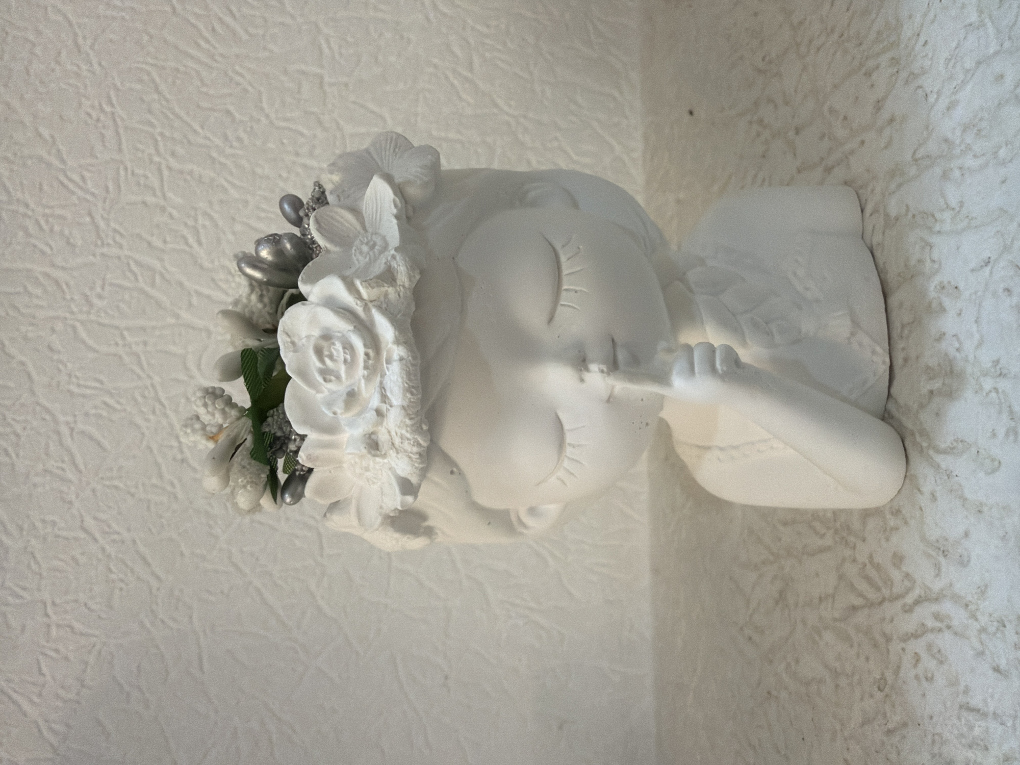 A white plaster bust of a girl with her eyes closed and finger to her lips, as if asking for quiet, sits on a textured surface. She wears a floral headdress adorned with white flowers and silver accents. The background is a textured, off-white wall. The scene is indoors.