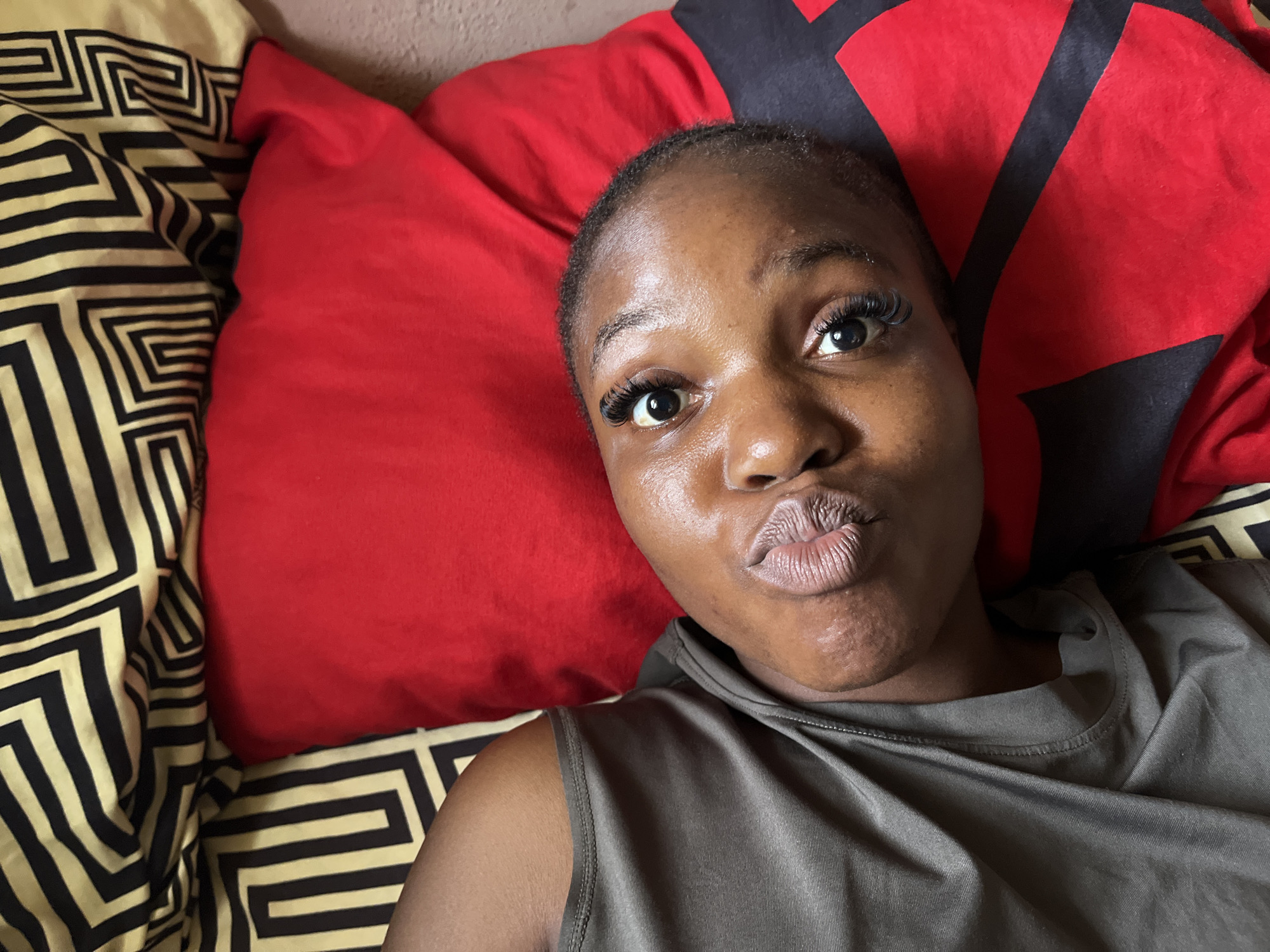 A young Black woman with long eyelashes and dark skin is lying on her back, looking up at the camera. She is wearing a grey tank top. The background consists of red and black pillows, and a patterned blanket in black and gold. The image was taken in Rafin Par, Nigeria.