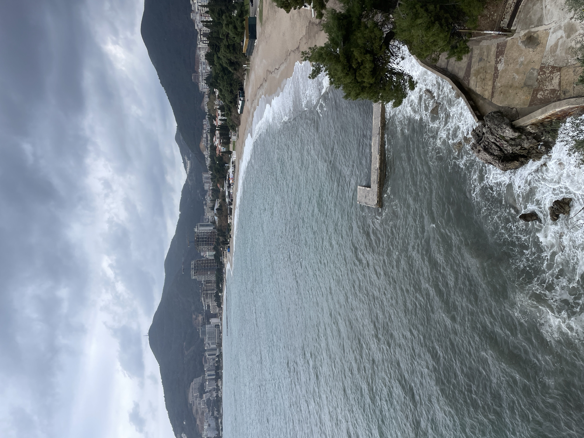 The image depicts a panoramic view of the coastline and urban area of Budva, Montenegro, under an overcast sky. The foreground features the turbulent, dark grey-blue waters of the Adriatic Sea, with white-capped waves crashing against a rocky shore and a concrete retaining wall that forms part of a curved pathway. A distinct, T-shaped concrete pier or jetty extends into the water from the lower-left quarter of the frame. Dense green coniferous trees and scrub vegetation line the immediate cliffside, with one prominent pine tree obscuring part of the pier.
Stretching across the midground is a wide, crescent-shaped sandy beach, likely Slovenska Plaza, where small waves break in a consistent white line. Several small, modular structures, including two dark green rectangular buildings and smaller white tents or kiosks, are visible on the sand. No individuals are present on the beach or in the water.
The background reveals the densely built urban landscape of Budva, characterized by numerous multi-story buildings, some appearing complete and others under construction, indicated by visible cranes. This urban development extends to the base of multiple large, dark green, tree-covered mountains that rise steeply behind the city. The most prominent mountain peak in the center-left background features a tall antenna or telecommunication mast at its summit. The sky is entirely covered by heavy, grey clouds, indicating a gloomy or inclement weather condition.