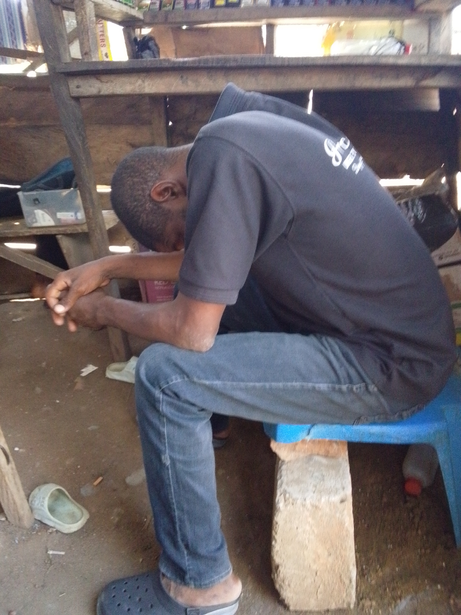An African man sits slumped forward on a blue plastic chair inside a makeshift wooden structure. He wears a dark t-shirt and jeans, with dark rubber sandals on his feet. His arms rest on his knees, his head bowed. A brick is used to support the chair. The floor is dirt. Another shoe sits on the floor to the left. The location is Lokoja, Nigeria.