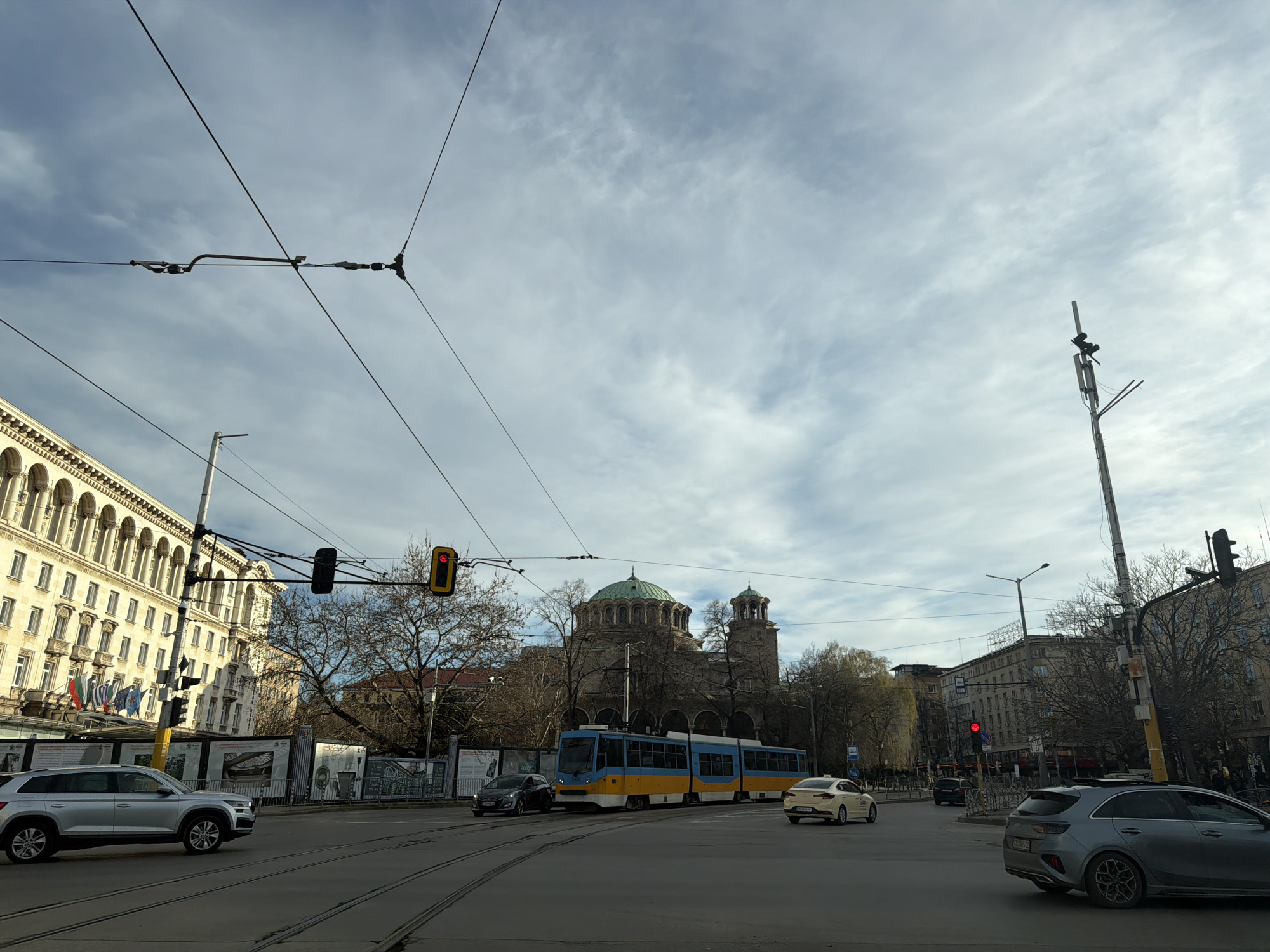 The image shows a street scene in Sofia, Bulgaria, with the St. Alexander Nevsky Cathedral visible in the background. A blue and yellow tram is crossing the street, and several cars are also present. The street is paved with tram tracks embedded in it. On the left side of the image, a building with many arched windows and balconies is visible, decorated with national flags. Traffic lights are present at the intersection. The sky is overcast. A cell tower is seen on the right side of the image.