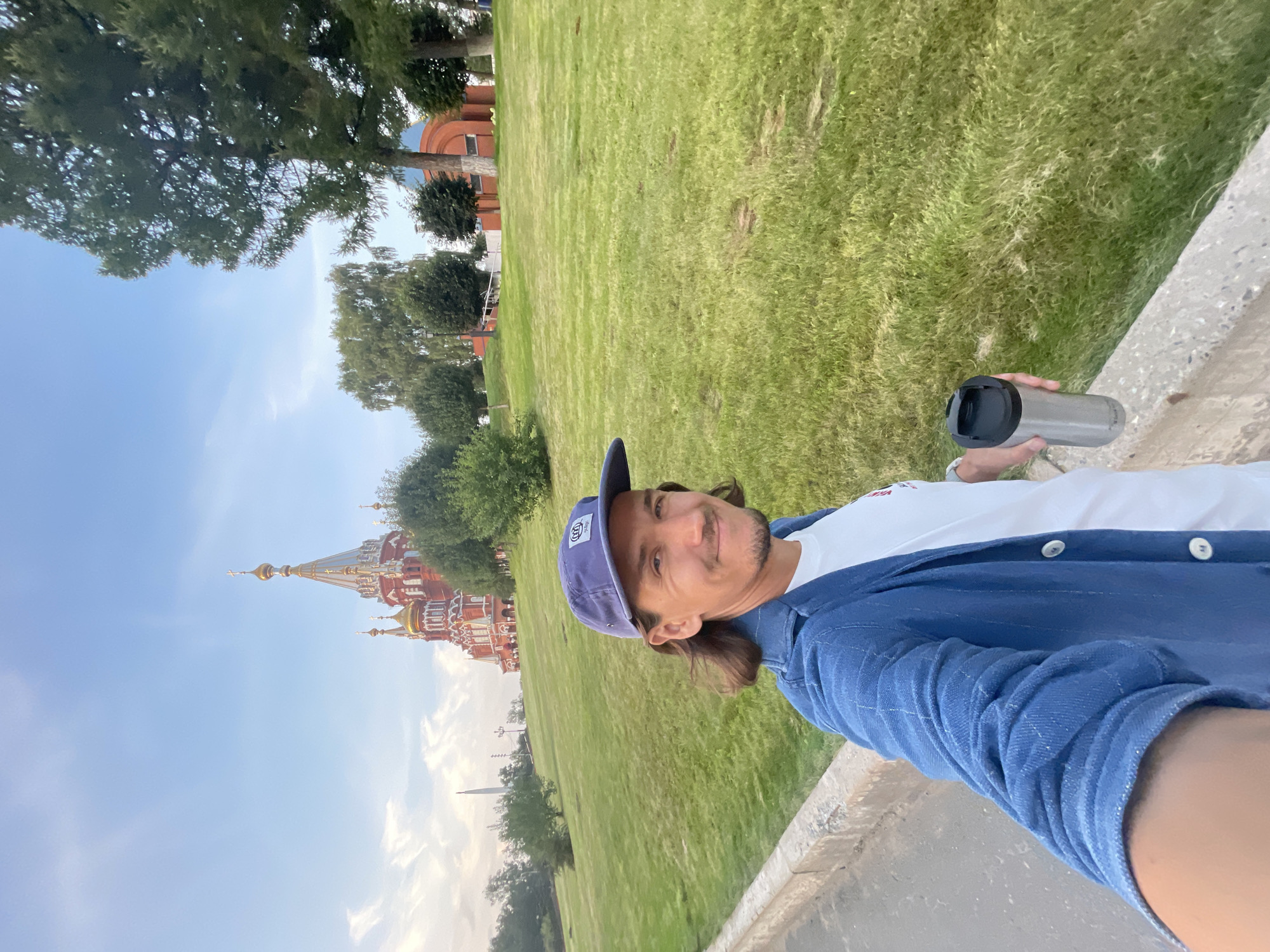 A man in a blue jacket and blue cap is taking a selfie outdoors. He is holding a silver travel mug in his right hand. Behind him is a large grassy hill, and in the distance, a red and gold cathedral with multiple domes is visible. The sky is cloudy and blue. The location is the Motherland Monument Park in Izhevsk, Russia, with the St. Michael's Cathedral in the background.
