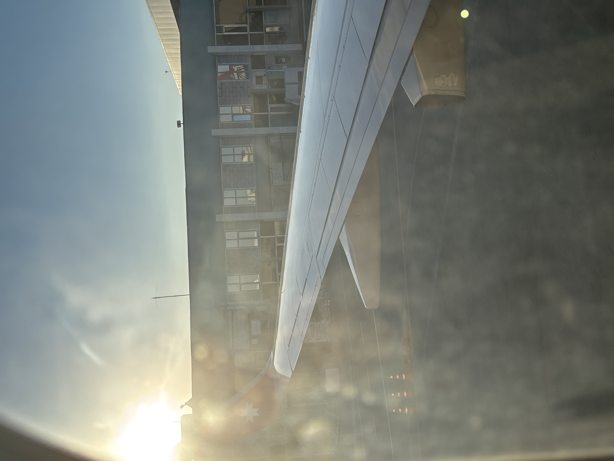 A view from inside an airplane shows a wing and a building outside the window. The building is multi-story and appears to be an apartment complex or office building with many windows. A flag is visible hanging from one of the windows. The wing of the plane is silver, and the aircraft is likely on the ground. The sun is low in the sky. The location is in the city of Новое Мартьяново, Russia.