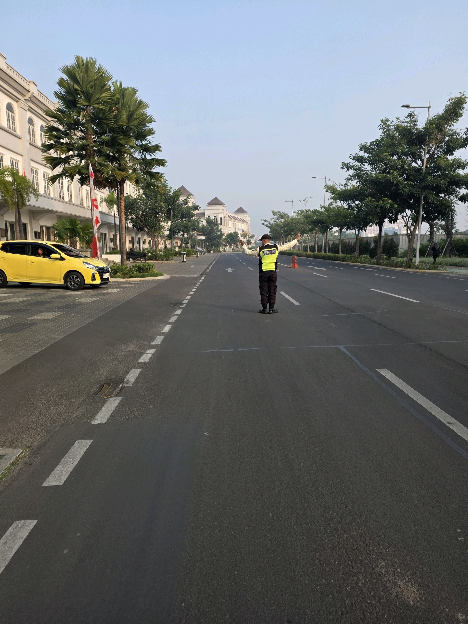 A lone traffic controller, wearing a high-visibility yellow vest and a dark cap, stands in the middle of a multi-lane asphalt road. Their arms are extended outwards, palms facing forward, in a clear gesture to stop traffic. The controller is positioned between white lane markings. The road stretches into the distance, lined by trees and modern, light-colored buildings on the left. A bright yellow car is parked on the left side of the road, near a pedestrian crossing marked with white stripes. The sky is clear and pale blue, indicating early morning or late afternoon. The scene appears to be in a developed urban or suburban area, possibly a resort or commercial district, given the architecture and manicured landscaping. The location is identified as Bojongsoang, Indonesia.