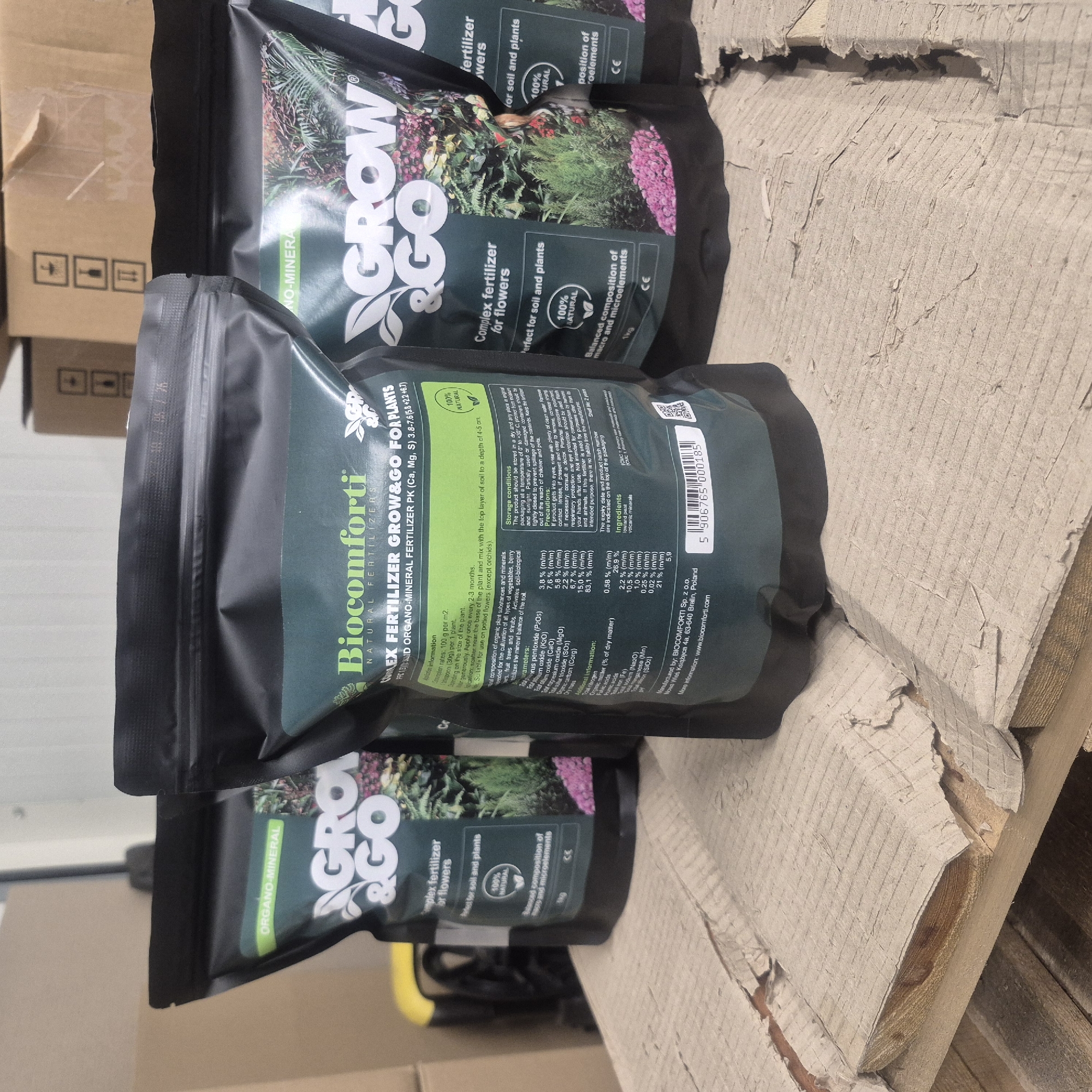 This image shows three black resealable bags of fertilizer, each with a green label. The bags are arranged on a wooden pallet covered with distressed cardboard. The bag in the foreground, labeled "Bioccomforti NATURAL FERTILIZERS COMPLEX FERTILIZER GROW&GO FOR PLANTS," is the most visible. Its label contains information about application, parameters, storage, precautions, and ingredients, listing components like potassium oxide, calcium oxide, and magnesium oxide. The bag also features a "100% NATURAL" seal and a barcode. To the left and right of the central bag are two other similar bags, one labeled "AGRO & GO" and the other partially obscured, also appearing to be fertilizer products. In the background, a yellow object and stacked cardboard boxes are visible, suggesting a storage or warehouse environment. The overall scene is indoors, likely a commercial or storage space. The text on the bag indicates that the manufacturer is BIOCCOMFORTI Sp. z o.o., located at Wąs Książęca 4a, 63-640 Bralin, Poland. The city of Nowa Wieś Książęca is near Bralin in Poland.