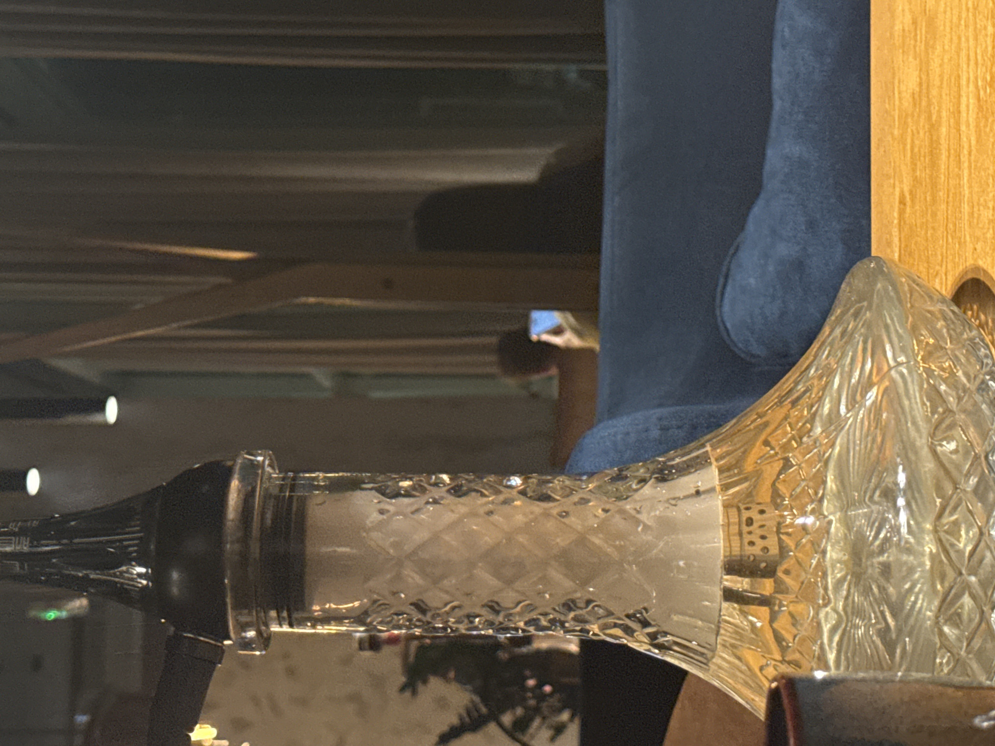 The image shows a close-up of a hookah with a glass base filled with clear liquid and some smoke. The hookah has a dark-colored top. In the background, out of focus, is a shadowy silhouette of a person holding a phone and looking at it, possibly in a dimly lit room with curtains. The scene appears to be indoors, possibly in a lounge or cafe. There is a blue sofa in the middle ground. The lighting is dim. There is no visible indication of the location being in Bratislava, Slovakia, or any specific city.