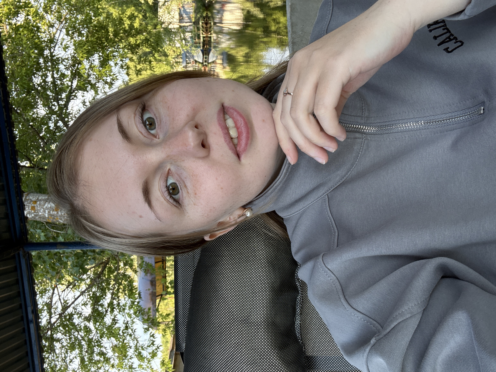 A medium-shot portrait captures a young woman from an elevated perspective, her head tilted ninety degrees counter-clockwise to the right. She has light skin with freckles and light brown hair that falls around her shoulders. Her green eyes look directly at the camera, and her lips are slightly parted, revealing her upper teeth. She wears a pearl stud earring in her right ear and a ring on the ring finger of her right hand, which is resting against her chin.
She is dressed in a grey quarter-zip sweatshirt, which has the partial text "CALI" (likely part of "CALIFORNIA") embroidered in black on the upper right chest area. The zipper of the sweatshirt is visible near her hand. She appears to be lying or leaning on a dark, finely woven mesh or textured surface, possibly an outdoor chair or lounge.
The background suggests an outdoor setting during daytime with bright sunlight. Green foliage from trees is prominent, and behind them, a body of water (possibly a lake or river) is visible, reflecting the bright sky and indistinct structures. Darker structures, possibly part of a canopy or railing, are also present in the foreground and mid-ground. The overall mood appears relaxed.