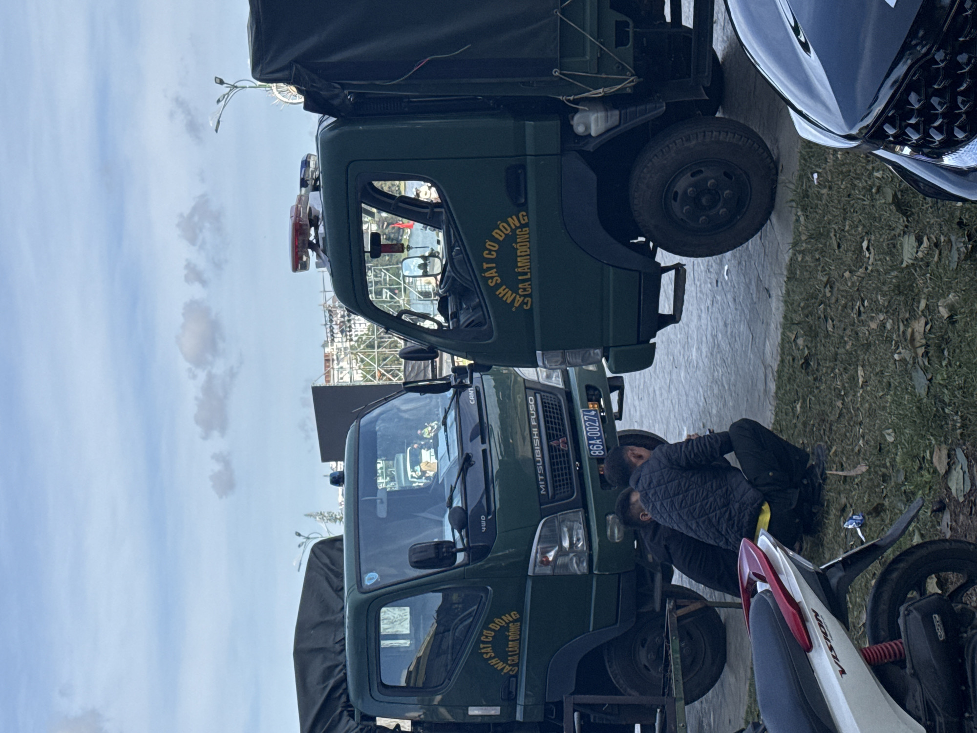 Two green military-style trucks with covered beds are parked side-by-side on a paved area adjacent to a grassy patch. The truck on the left has yellow lettering on its door that reads "CẢNH SÁT CƠ ĐỘNG CA LÂM ĐỒNG", and its license plate is "86A-00274". The truck on the right also has yellow lettering on its door with the same text. Both trucks have emergency lights on their roofs. Three men are present; two are sitting or crouching on the ground between the trucks, and one is visible inside the cab of the left truck. A white and red scooter is parked in the foreground to the left, and the front of a black car is visible in the foreground to the right. The background shows buildings and trees, suggesting an urban or semi-urban setting. The sky is cloudy. The scene appears to be located in Da Lat, Vietnam, as indicated by the text on the trucks.