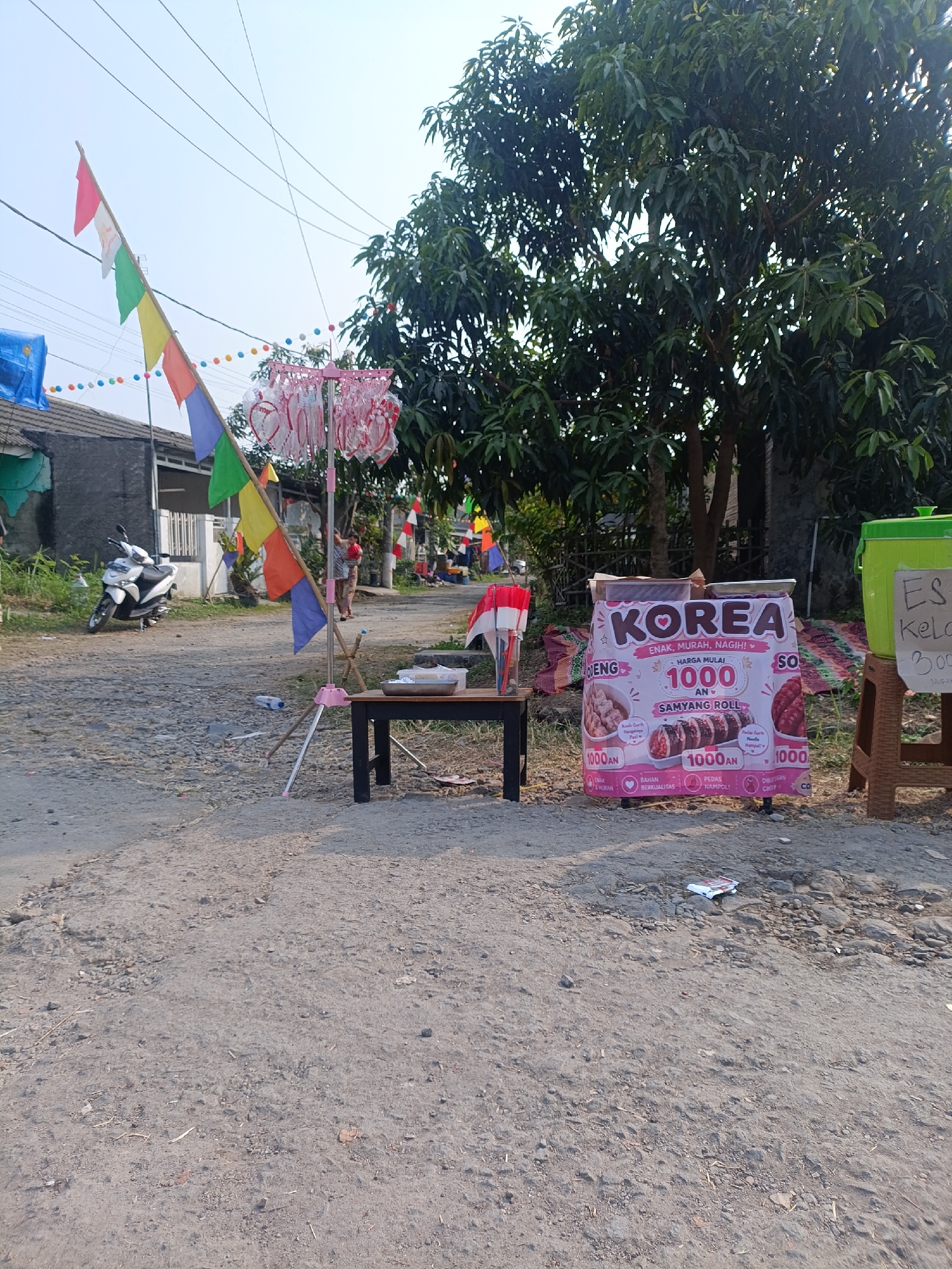 The image depicts an outdoor scene in the city of Cariu, Indonesia, likely on a street or along a path. In the foreground, a rough, gravelly surface suggests a rural or less developed area. A black table with a metal tray sits on the right side of the image.
Dominating the center of the frame is a vibrant pink banner advertising "KOREA" food products, specifically "SAMYANG ROLL," with prices indicated as "HARGA MULAI 1000 AN." The banner features images of food items and text in Indonesian. To the right of the banner, a brown stool holds a green cooler.
The background reveals a more developed area with houses, some with a dark gray facade. A white scooter is parked to the left, and a series of colorful flags, including red and white Indonesian flags, are strung across wires overhead, indicating a possible festive occasion or market. Further in the background, a large, leafy tree stands prominently. Two individuals are visible in the middle ground, one standing and the other partially obscured. The overall impression is of a temporary street-side vending setup in a lively local setting.