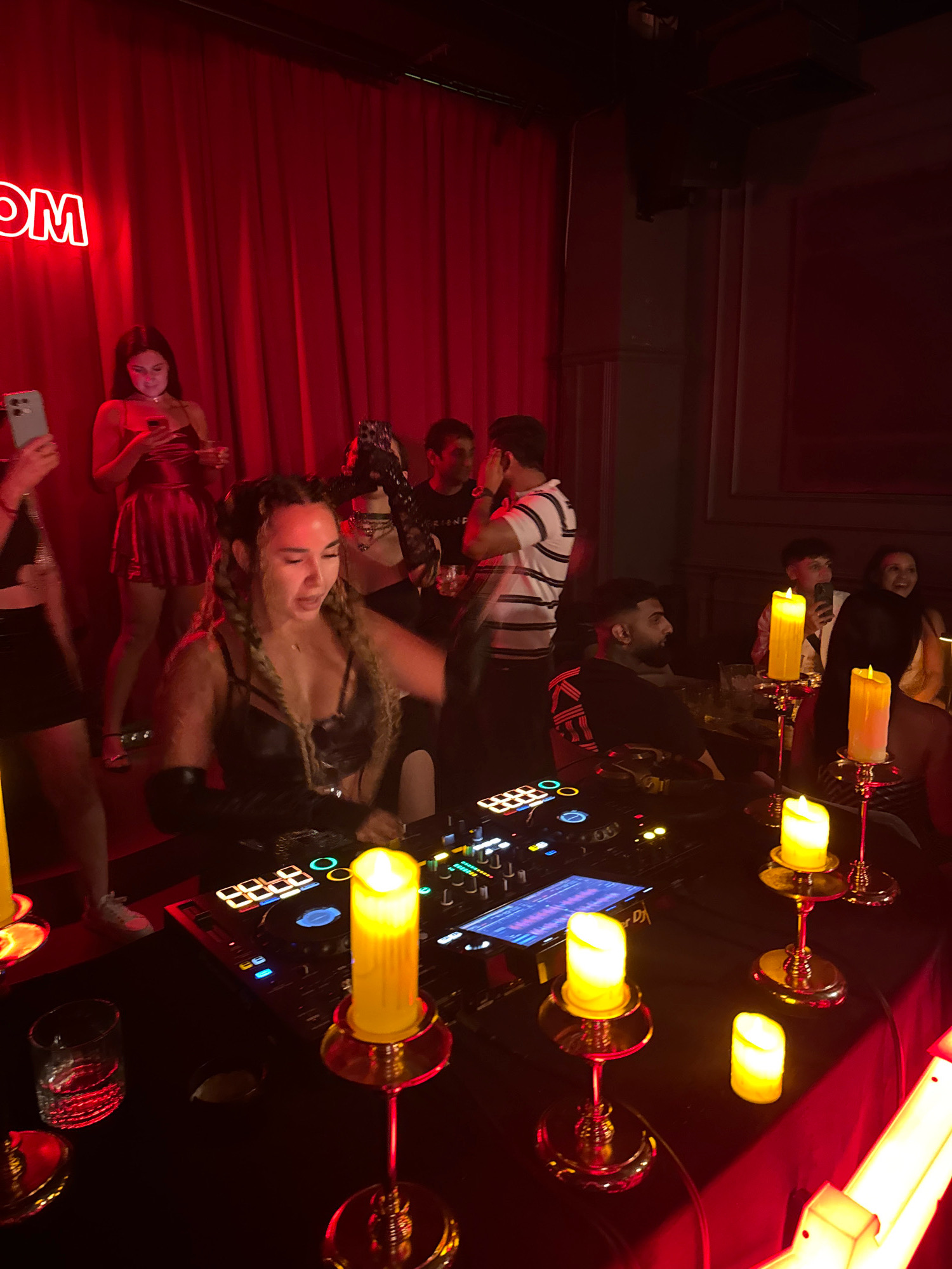 In a dimly lit interior, likely a nightclub or event space in North Kuta, Indonesia, a group of people are engaged in various activities under red ambient lighting. In the foreground, a woman with braided hair, wearing a black outfit with cutouts and gloves, is actively operating a DJ console. Her mouth is open, suggesting she is speaking or singing. The console displays a screen with a waveform. Nearby, on a table covered with a black cloth, are several decorative LED candles, three of which are lit, casting a warm glow. A glass with a clear liquid sits to the left of the DJ console.
In the mid-ground, a woman in a red dress and heels is holding a phone. To her left, another person, partially obscured, is also holding a phone. Behind them, a man in a striped t-shirt appears to be looking away from the camera. Further back, another man is visible, and to his right, a woman is smiling. The background features red curtains and a neon sign that partially reads "OM". The overall atmosphere suggests a lively social gathering or performance.