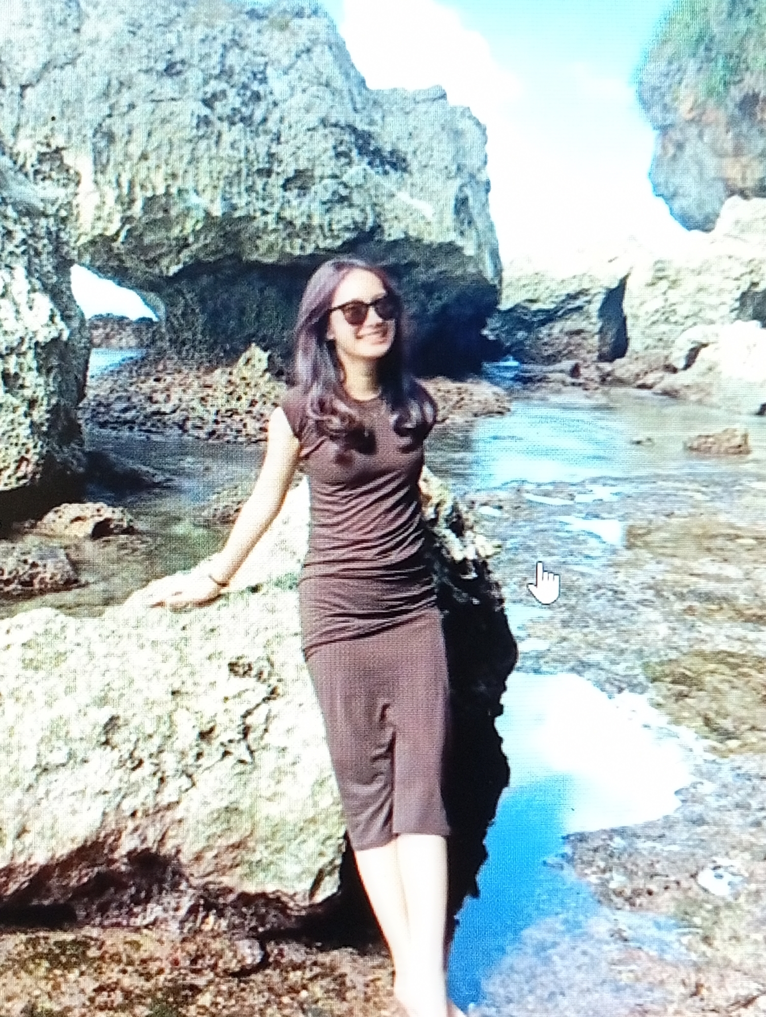 A woman with dark hair and sunglasses stands on rocks by the sea. She is wearing a brown, sleeveless, knee-length dress. The background features large rock formations and calm water, suggesting a coastal location. The scene appears to be outdoors, with a bright sky visible above the rocks.