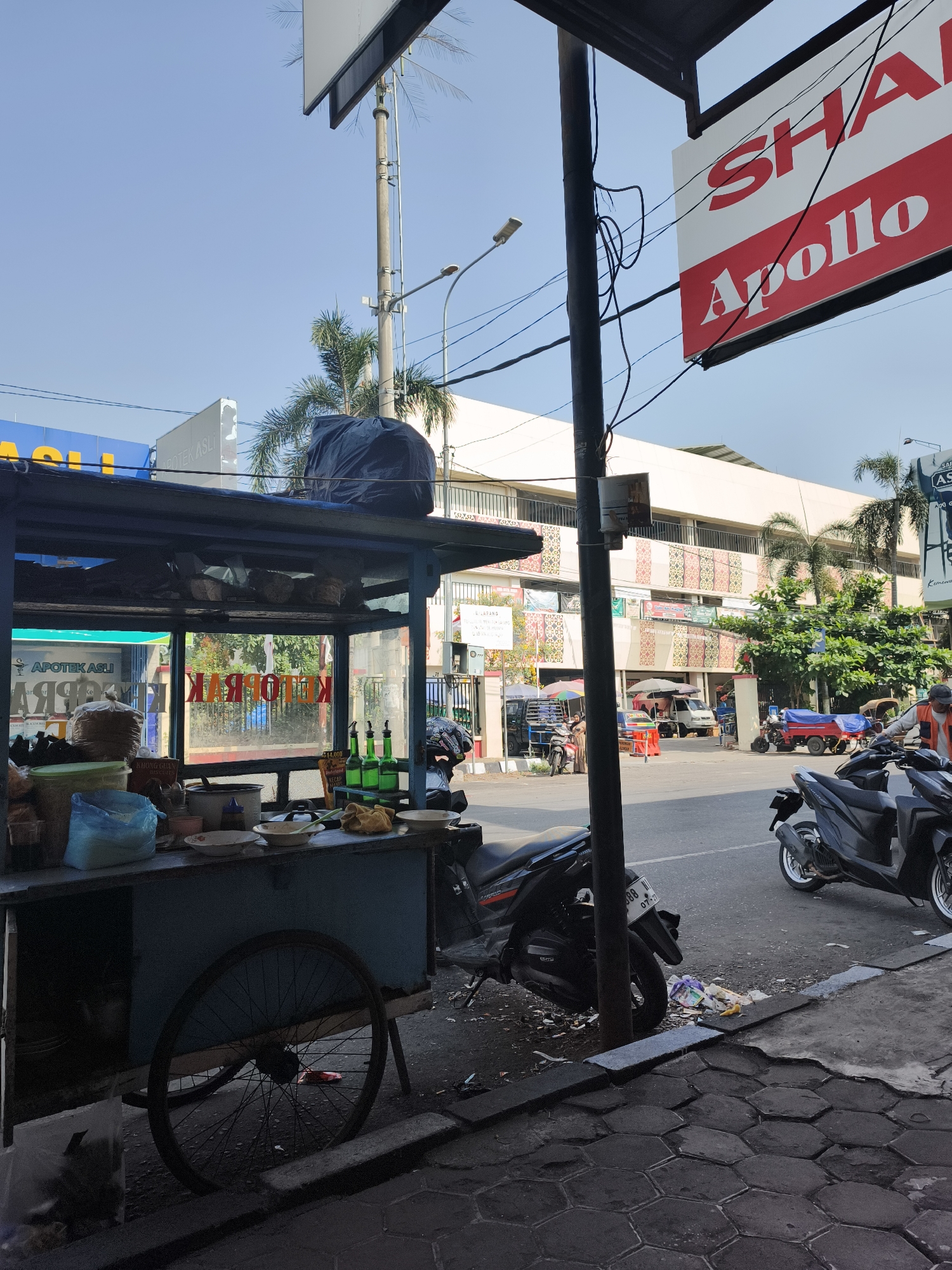 The image depicts a street scene in Pekalongan, Indonesia, under a clear blue sky. In the foreground, a food cart with a blue frame and a large wheel is positioned on a tiled sidewalk. The cart displays various items, including bowls, bottles, and bags of goods. A black tarp covers a large object on top of the cart. To the right of the cart, several motorcycles are parked on the street.
Behind the cart, a building with a white facade and decorative patterns can be seen. A large red and white sign with "SHARP Apollo" written on it hangs from a pole. Across the street, more buildings and various vehicles, including motorcycles and a small red truck, are visible. A few people are present, some appearing to be vendors or customers. The environment suggests a busy commercial area with food vendors and shops. The overall impression is that of a typical day in an Indonesian city.