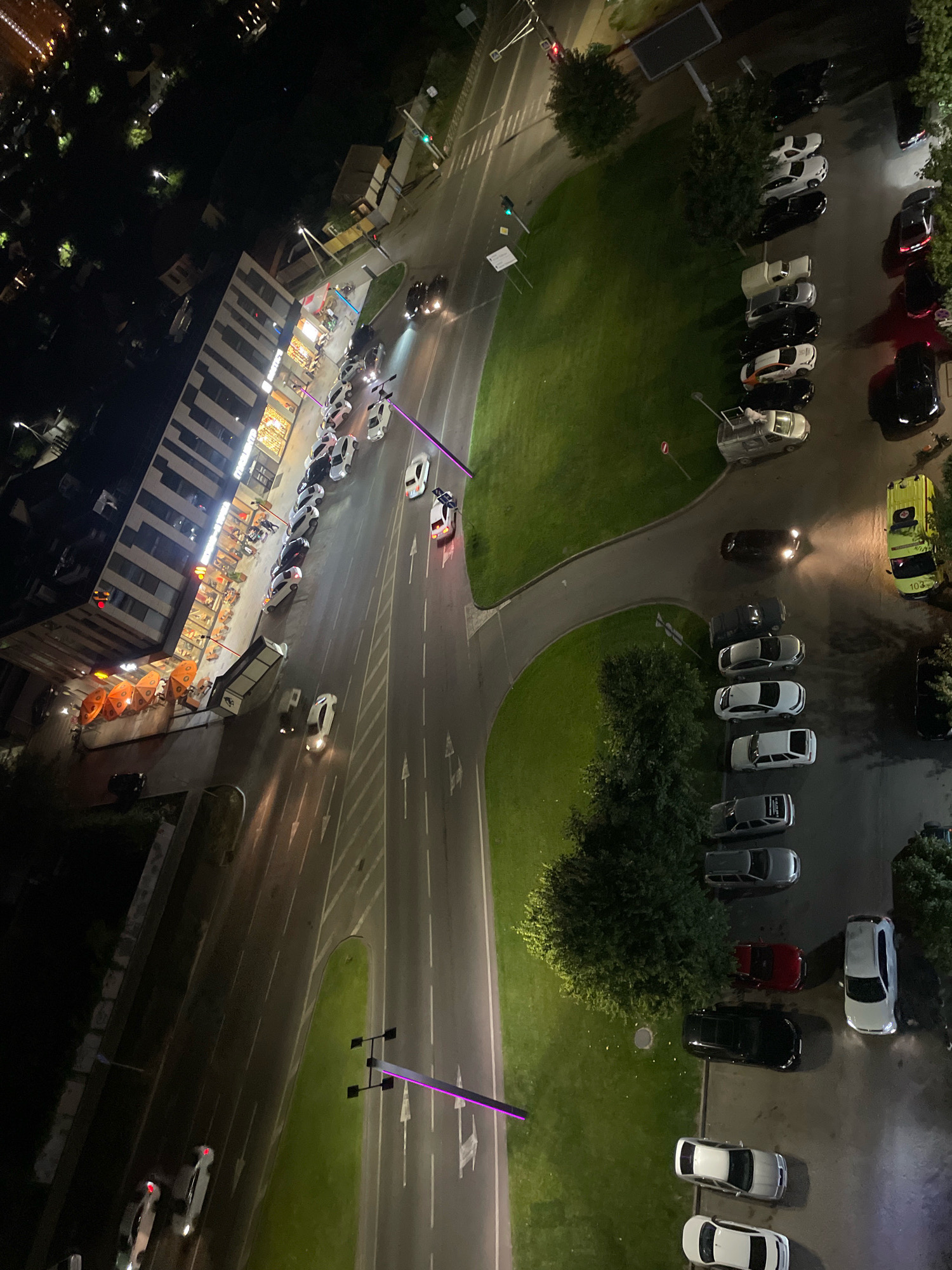 The image shows an aerial view of a street in Krasnodar, Russia, at night. A building with illuminated windows is on the left, adjacent to a road with parked cars and moving vehicles. A green grassy area with trees is on the right. Several cars are parked along the street and in parking lots. One vehicle, possibly a food truck or service vehicle, is yellow with the number 103 on its side. Purple illuminated structures resembling lights or art installations are present on the roadside. The overall scene depicts a nocturnal urban environment with vehicular and pedestrian activity.