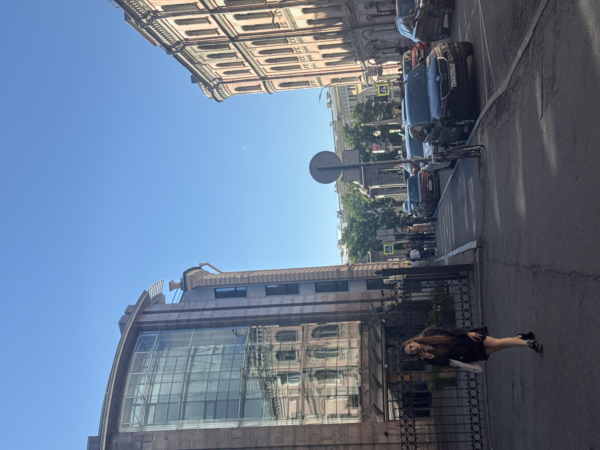 The image depicts a street scene in Saint Petersburg, Russia. On the left side, a building with a modern, curved glass facade dominates the foreground, reflecting an older, classical building across the street. This older building features multiple stories with arched windows and ornate detailing. The sky above is a clear, bright blue, suggesting a sunny day.
The street is lined with parked cars, including a black Skoda with the license plate "M197KG 198". Further down the street, other vehicles and pedestrians can be seen. A traffic sign with a yellow diamond and a black symbol is visible, as well as a circular sign on a pole. Trees line the opposite side of the street, adding greenery to the urban landscape.
In the foreground, on the right side of the image, a young woman with long, light brown hair is walking along the sidewalk. She is wearing a dark-colored outfit, possibly a dress or a skirt and top, and sandals. She is looking towards the camera and appears to be speaking on her phone. Her expression is neutral or slightly smiling. A white bag is hanging from her shoulder. A decorative black metal fence runs along the sidewalk in front of the modern building.
The time of day appears to be daytime, given the bright sunlight and clear sky. The overall atmosphere seems to be a typical urban environment on a pleasant day.