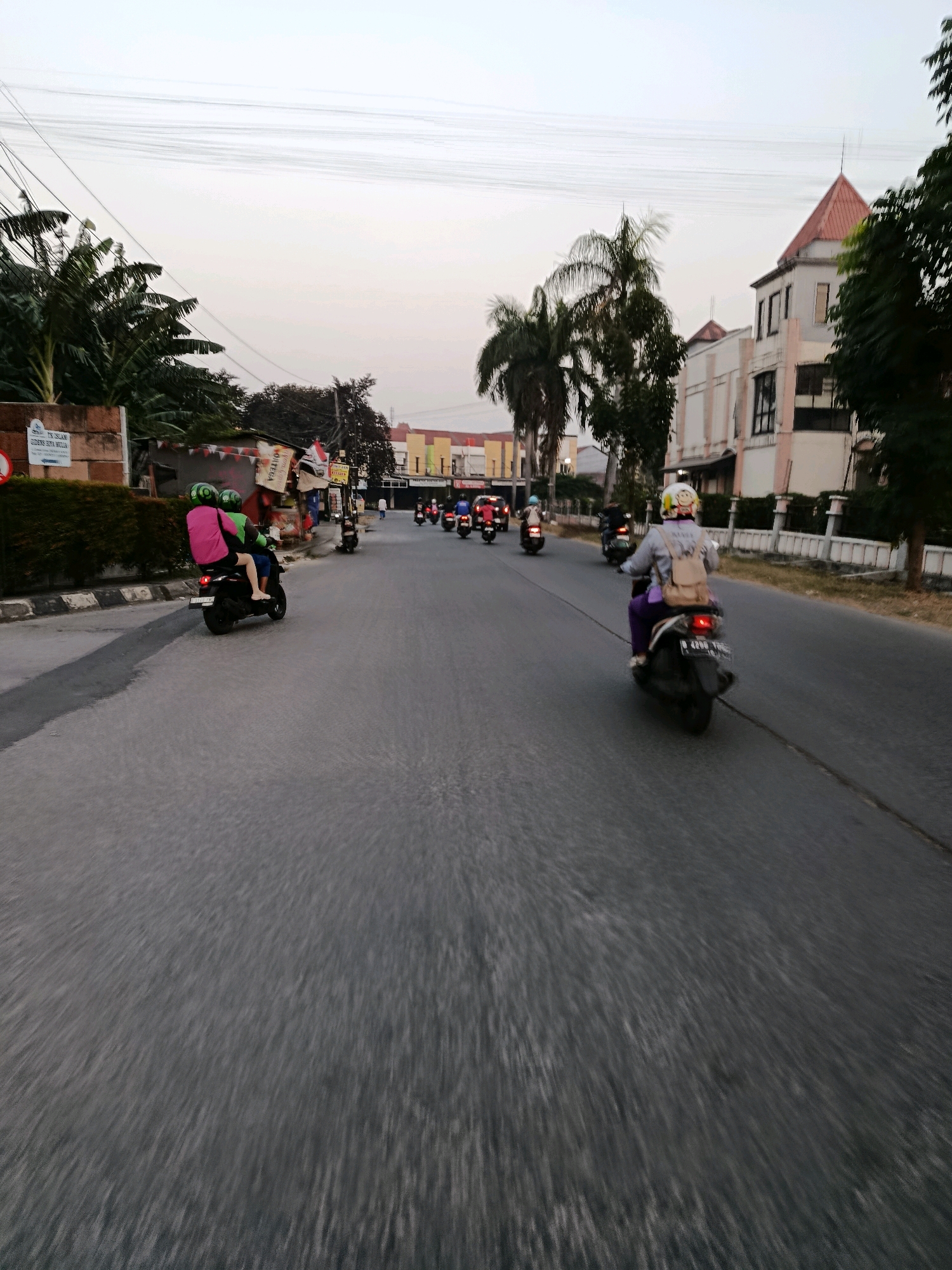 In Bekasi, Indonesia, a street scene unfolds with a line of motorcycles navigating the asphalt road under an overcast sky. In the foreground, a scooter carrying two individuals, one wearing a neon green helmet and the other a pink jacket, moves away from the viewer. To the right, another scooter rider, adorned with a yellow helmet featuring a smiley face and a beige backpack, also travels forward. Further down the road, a procession of motorcycles stretches into the distance, their forms blurred by motion. Flanking the road are buildings with a pale facade and a prominent reddish-brown roof on the right, alongside lush green foliage and palm trees. Signs and structures are visible along the left side of the road, suggesting an urban commercial or residential area. The overall ambiance is that of a typical day on a moderately busy street in an Indonesian city.