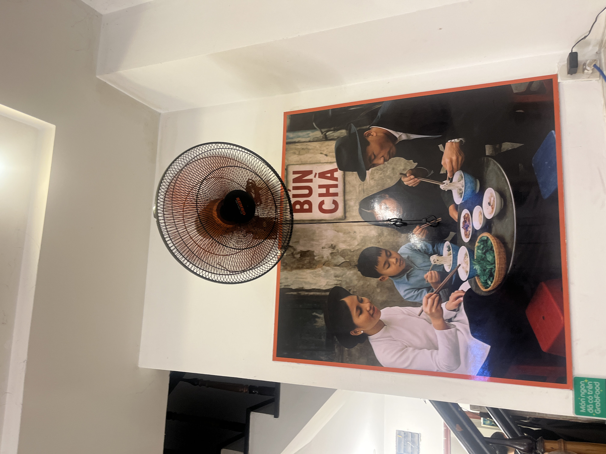 The image shows a framed photograph on a white wall of a restaurant. The photograph depicts a family eating a meal of noodles. A woman in traditional Vietnamese clothing, a young boy, and a man in a suit and hat are seated around a table. The man in the suit is holding chopsticks and eating noodles. The photograph has "BUN CHA" written in red letters on a white background in the upper right quadrant of the image. A ceiling fan is mounted on the wall above the photograph. A green sign with Vietnamese text and the GrabFood logo is visible in the lower left corner of the image. The environment appears to be a casual dining establishment. The location of this establishment is in Nha Trang Ward, Vietnam.