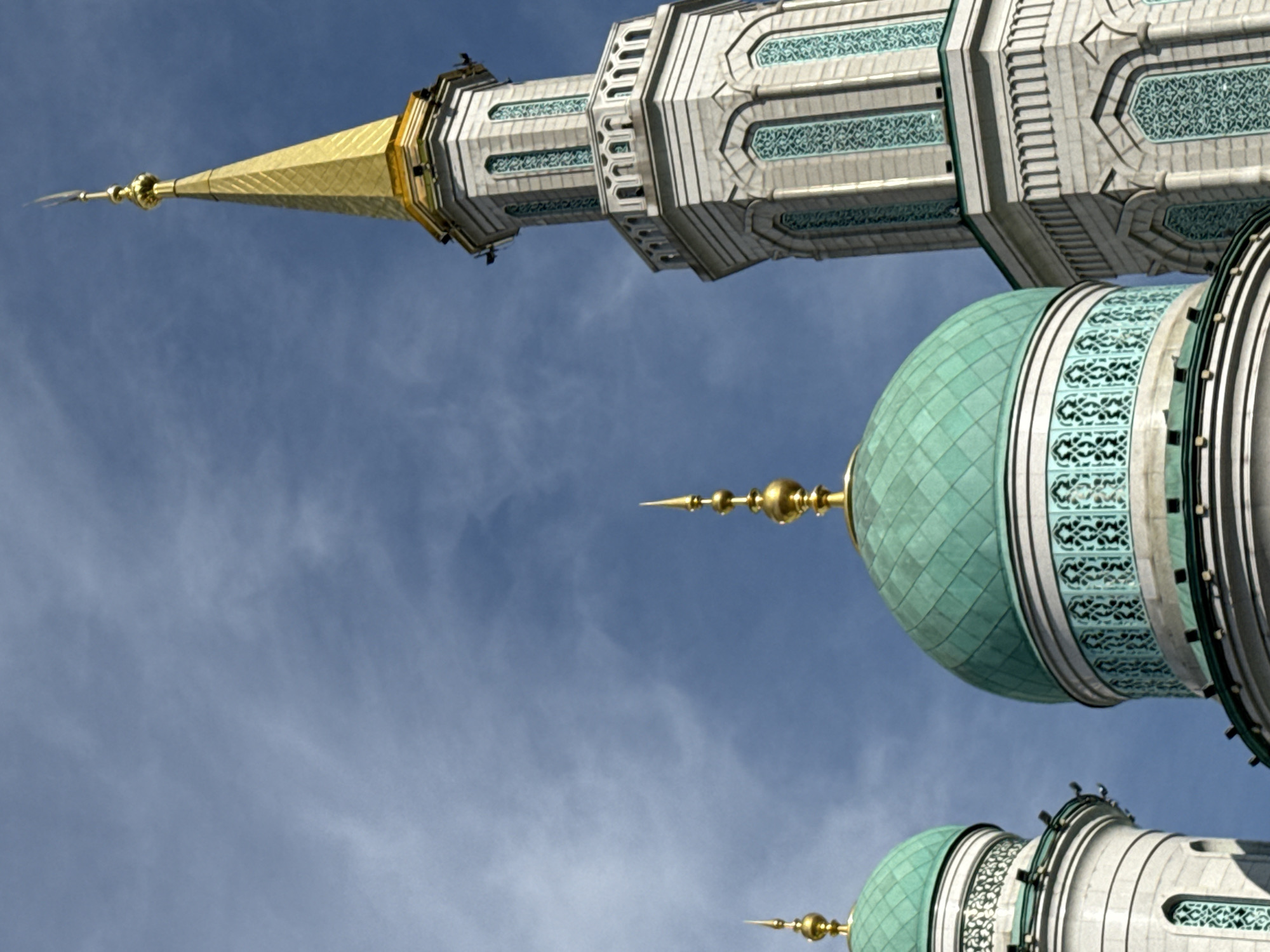 This image shows a close-up, low-angle view of the Moscow Cathedral Mosque in Moscow, Russia, against a cloudy blue sky. The scene is dominated by the mosque's architecture, featuring two prominent green domes and a tall, ornate minaret topped with a golden spire. The domes are ribbed and capped with golden finials. The minaret is octagonal, constructed of light-colored stone, with decorative teal accents and arched openings near its top. The overall impression is one of grandeur and religious significance. No people or animals are visible in the frame.