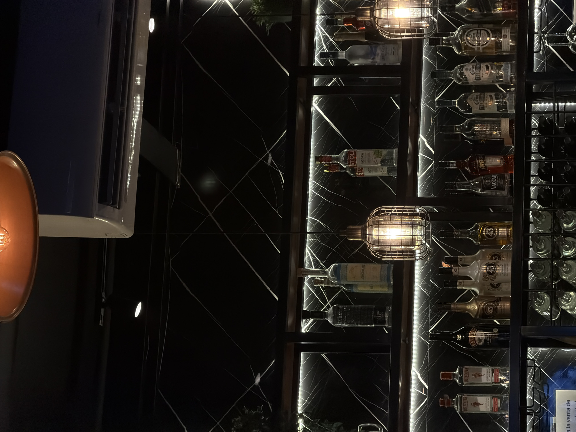Here's a description of the image:
The image shows a well-stocked bar with bottles displayed on shelves against a black marble-like background featuring white vein patterns. Bottles of various alcoholic beverages, including vodka (Stolichnaya), gin (Beefeater), and liqueur (Licor 43, Baileys), are visible. Light fixtures with a caged design are positioned among the shelves, casting a warm glow. The setting appears to be an upscale bar or restaurant with a modern design aesthetic. The OCR suggests that this could be "San Bernardino Tlaxcalancingo, Mexico".
