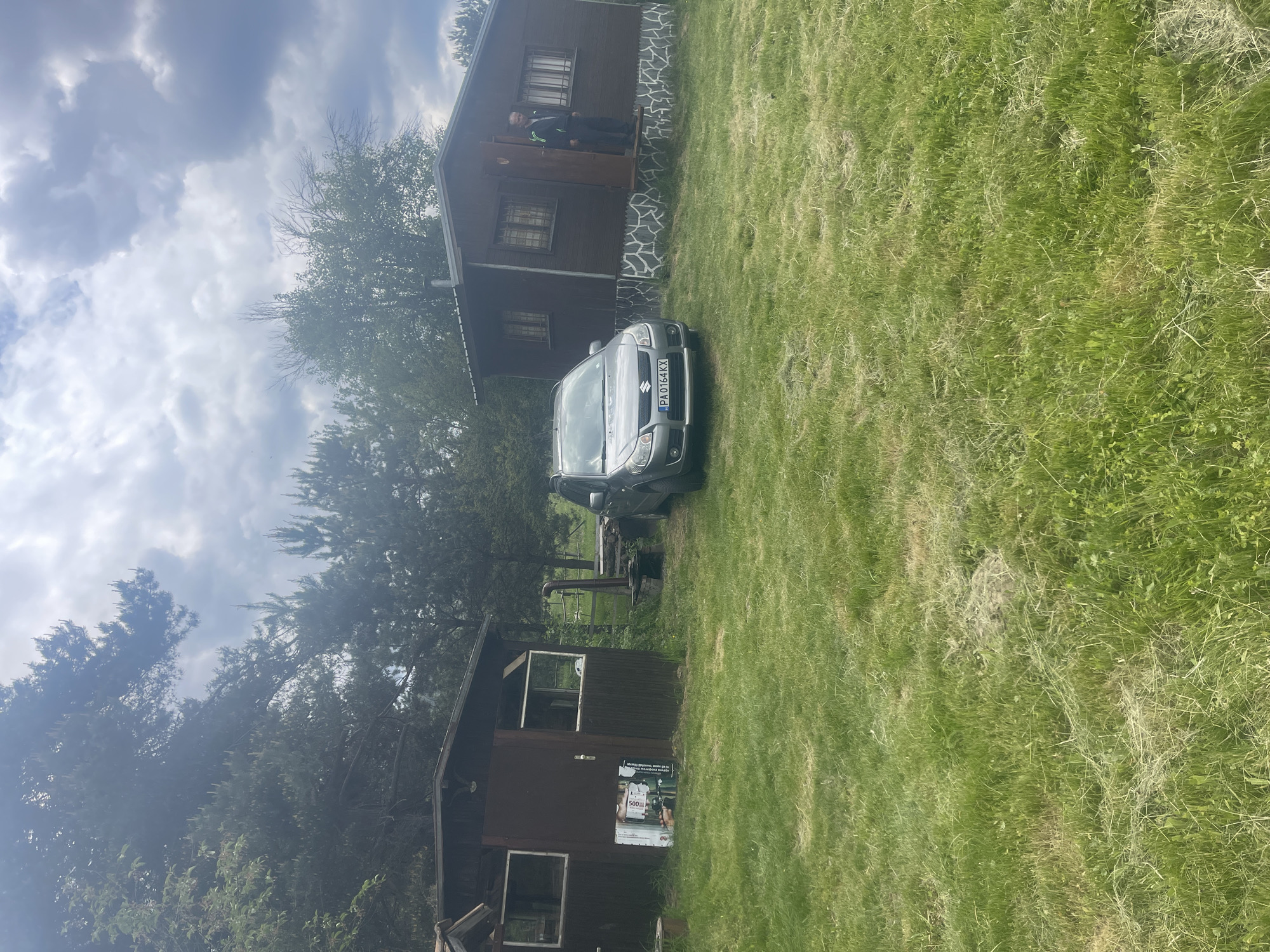 The scene is in Batak, Bulgaria. It depicts a gray Suzuki SX4 parked between two wooden buildings. The building on the left is a small shed with a poster affixed to it, while the one on the right is a cabin with a person standing in the doorway. The environment consists primarily of a grassy lawn under a cloudy sky.
