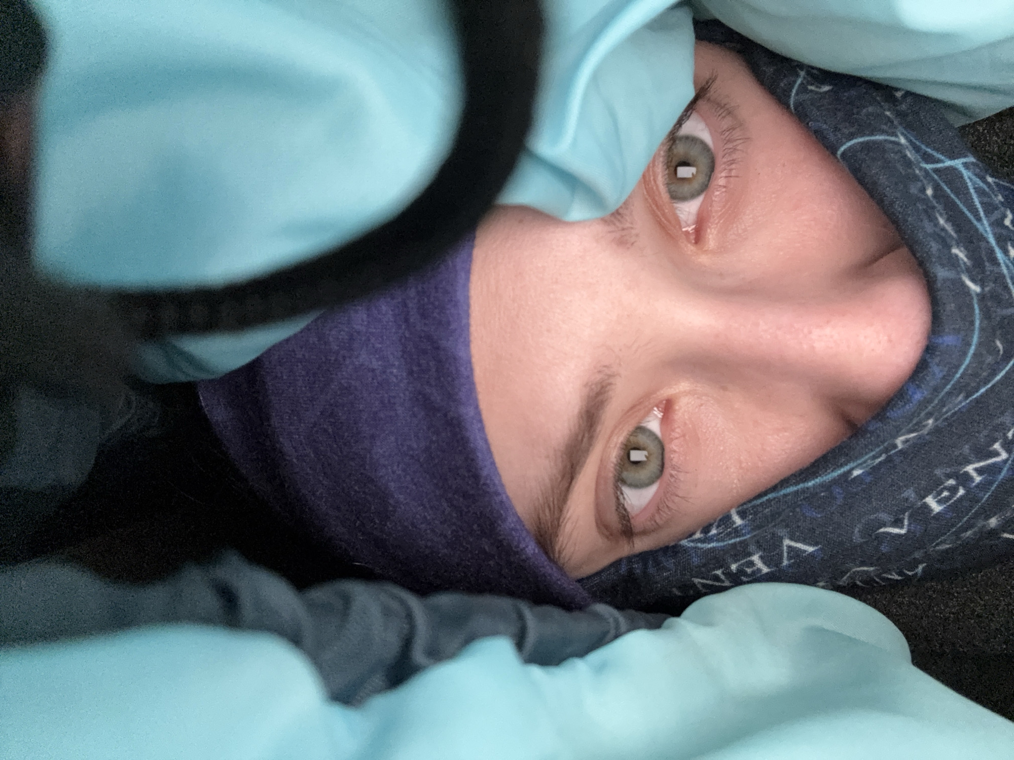 A close-up, angled shot captures a person's face, primarily their eyes, nose, and forehead, from an overhead perspective. The individual is heavily bundled in multiple layers of fabric, suggesting a need for warmth. Light blue material, resembling a sleeping bag or thick blanket, frames the face from the left and bottom. Dark blue or purple fabric, possibly a beanie or balaclava, covers the sides of the face and forehead. On the right side of the face, a dark blue patterned fabric, likely a neck gaiter or hood, is visible, featuring partial white text that appears to spell "VEN" and "NVA," possibly part of "VENT" or a similar word, partially obscured or inverted due to the fabric's folds.
The person's eyes are open and alert, looking slightly upward and outward. Their irises are light in color, possibly green or hazel, and show distinct rectangular reflections, suggesting an artificial light source or a window nearby. The expression is neutral and observant. A dark, blurry, circular object, potentially a strap or part of some equipment, is visible in the upper left foreground. The tight bundling indicates an environment where warmth is essential, consistent with potentially cold conditions in Lesnoye, Russia. The specific time of day or outdoor weather cannot be precisely determined, but the scene conveys a sense of being protected from the cold while resting or observing.
