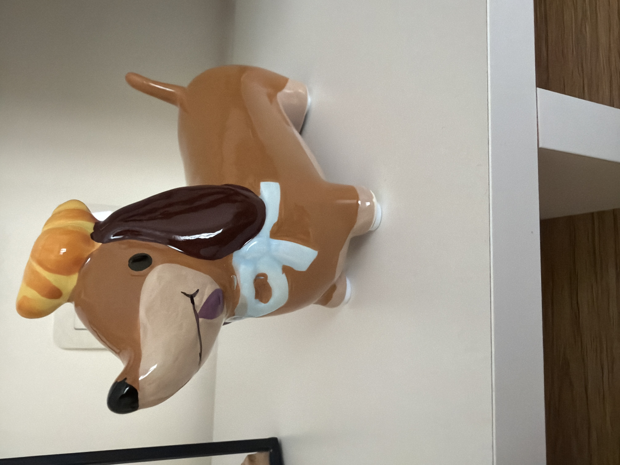 A ceramic dachshund figurine, predominantly brown with a light brown snout and purple tongue, is positioned on a white shelf. The figurine wears a blue bow around its neck and a croissant-like hat with yellow and orange stripes on its head. The background consists of a plain white wall with a light switch visible. The shelf is supported by white legs, and a wooden floor is visible beneath. The scene is indoors, with no specific geographical location in Pazardzhik, Bulgaria, identifiable from the image.