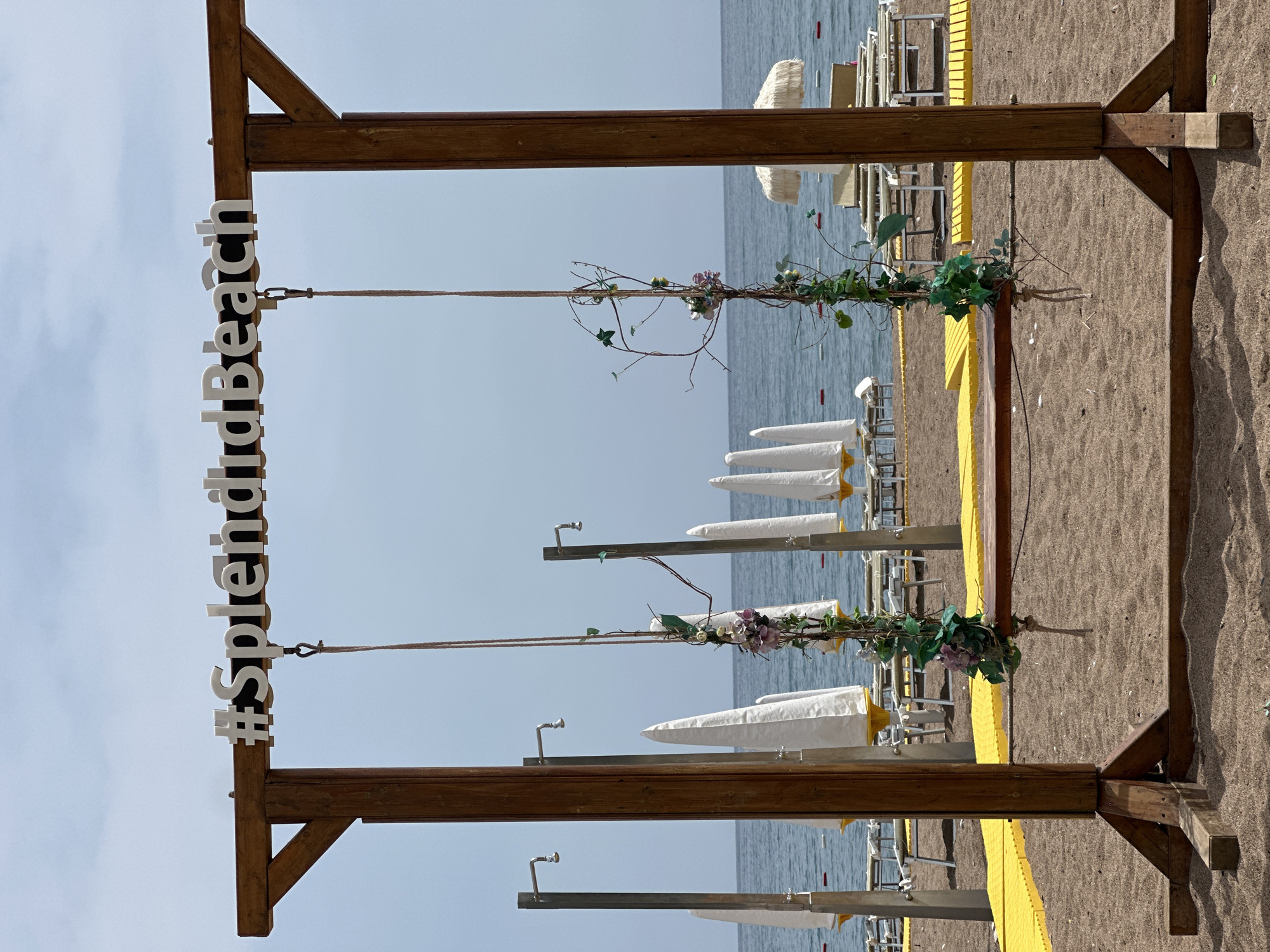 The image depicts a beach scene in Boreti, Montenegro. A wooden frame stands on the sandy beach, with the hashtag "#SplendidBeach" attached to the top left corner. A rope is strung across the frame, adorned with foliage and purple flowers. In the background, the sea extends to the horizon. Beach umbrellas and chairs are arranged along the shore. The ground is sandy, and a yellow barrier separates the beach area from the background.