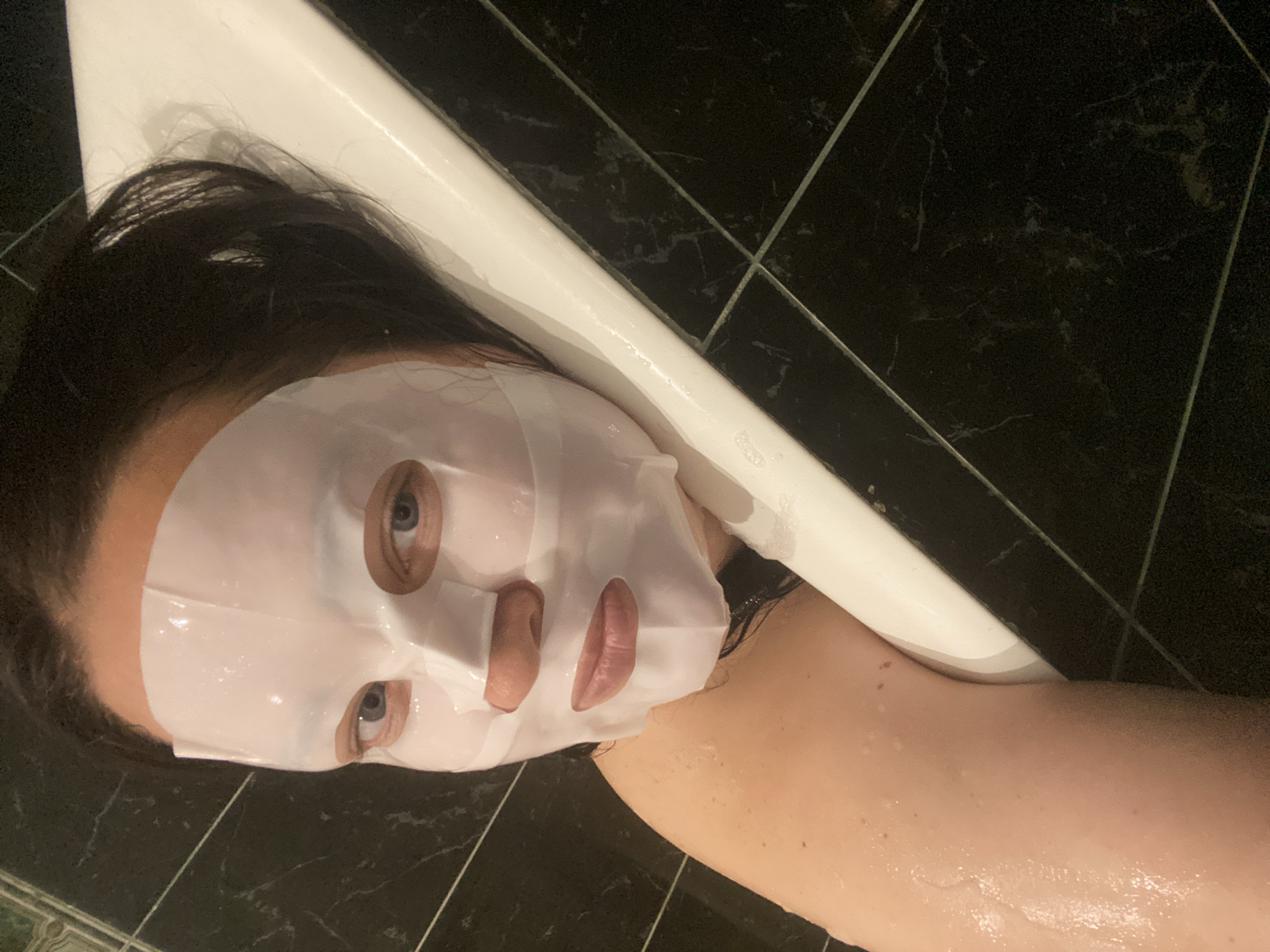 A person wearing a sheet mask lies near the edge of a white bathtub. The background consists of black tile with light-colored grout. There is no information to indicate this image was taken in Sochi, Russia.