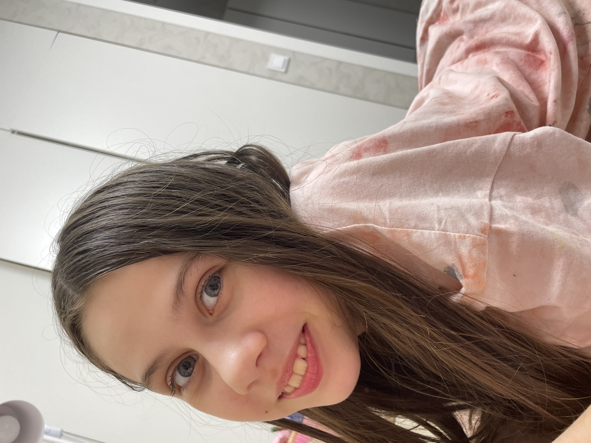 A young woman with long brown hair and blue eyes is smiling at the camera. She is wearing a pink hooded sweatshirt with what appears to be paint stains on it. The background is a simple white wall with a light switch. The location cannot be determined to be Izhevsk, Russia.