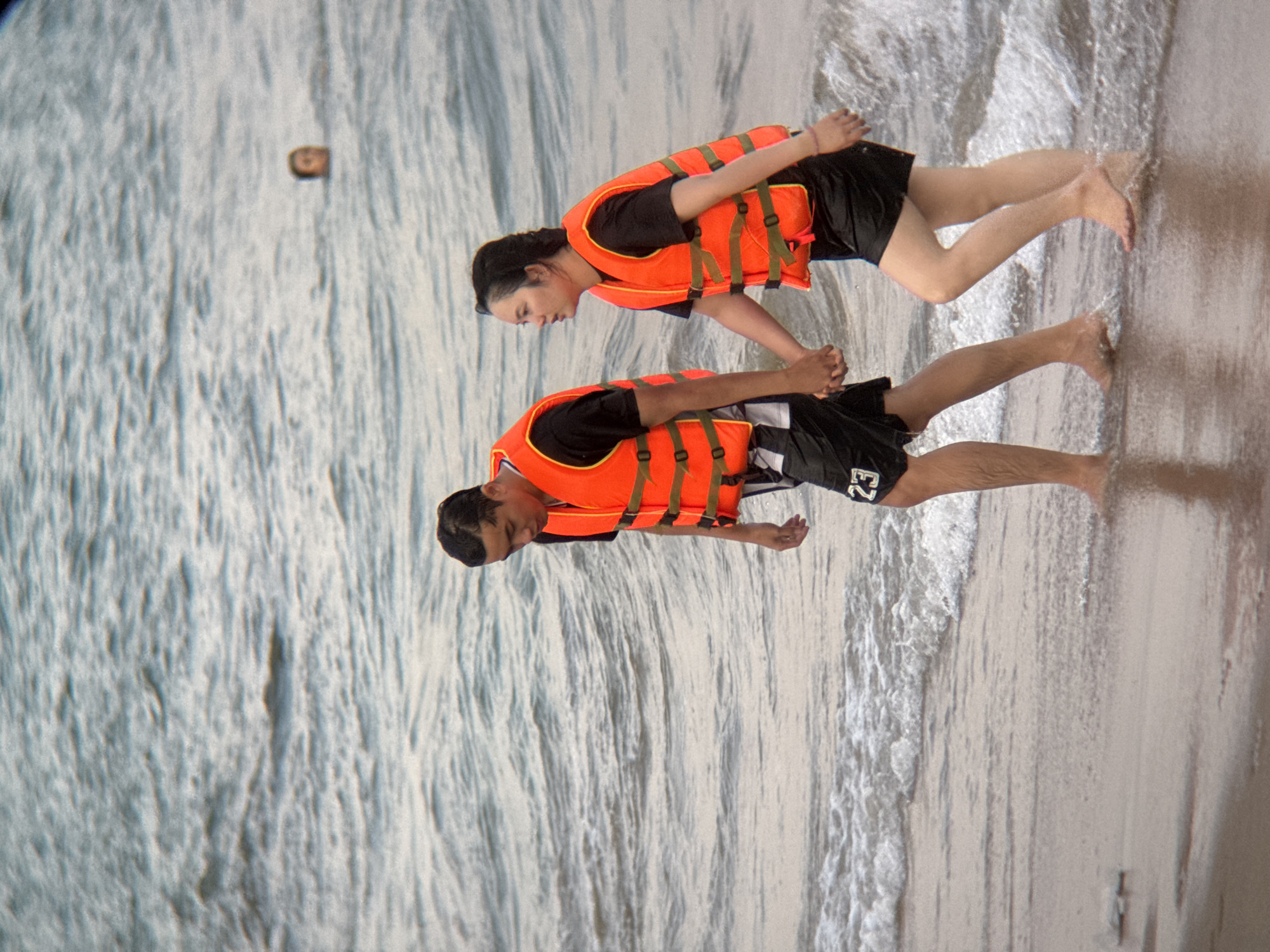 An adult couple walks hand-in-hand along a sandy beach. The man, on the left, wears a black t-shirt and black shorts with a white number "23" on the left thigh, and an orange life vest. He is looking down. The woman, on the right, wears a black t-shirt and black shorts, and an orange life vest. She is looking forward and to her left. They are walking towards the right of the frame. Gentle waves lap at their feet. In the background, a person's head is visible above the water, further out to sea. The location is a beach in Nha Trang, Vietnam.