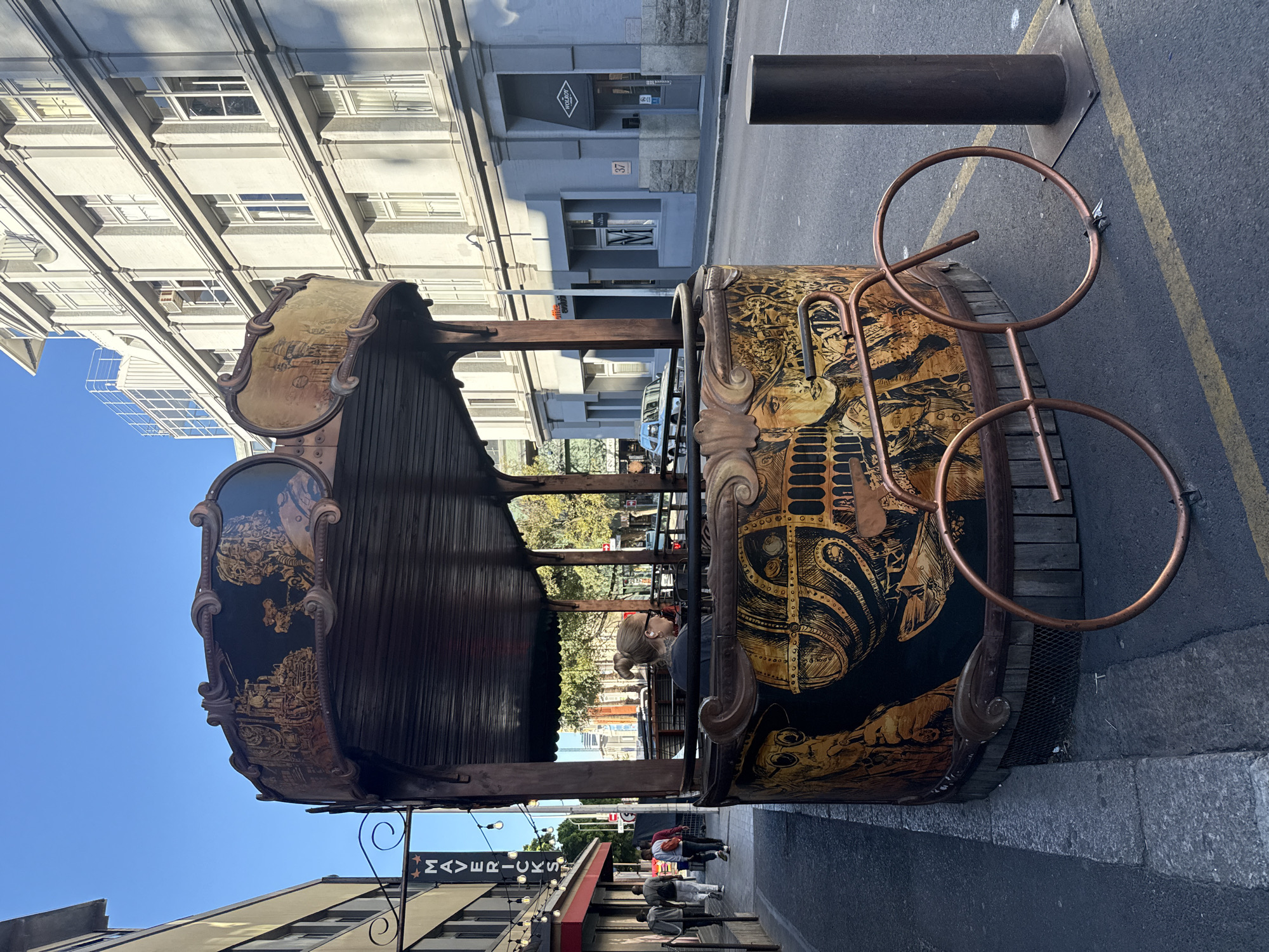 The image depicts an ornate, vintage-style street installation, possibly a seating structure or decorative art piece, located on a city street. The structure features detailed artwork with gold and black hues, mounted on a wooden base with copper accents. Adjacent to the structure, a dark metal pole stands near the edge of the street. In the background, buildings with multiple floors, and numerous windows are visible. Street signs and a business front labeled "Mavericks" are visible on one side of the road. On the opposite side, a building with "37" sign and "WolRoy" signboard above the entrance. People are present in the background, walking and seated nearby. Based on architectural style and street signs, the location is possibly in Cape Town, South Africa.