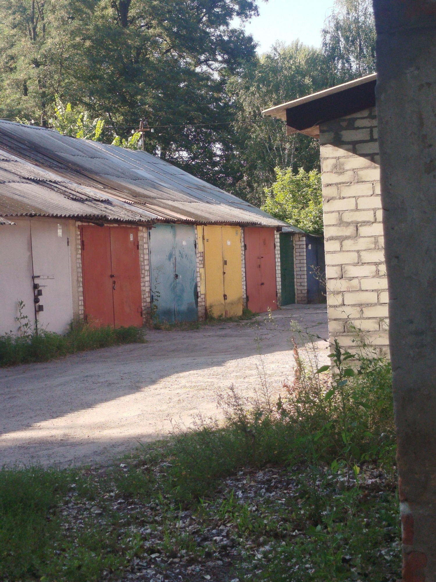 A line of multi-colored garage doors, some weathered and with visible rust, are set into a long, low building with a corrugated metal roof. The garages are painted in various colors, including white, orange, blue, and yellow. The ground in front of the garages is a dirt and gravel road, partially shaded by dappled sunlight. Overgrown grass and weeds grow along the base of the garages and on the edges of the road. In the background, lush green trees form a dense canopy. A brick structure, partially visible on the right side of the frame, suggests further construction or an adjacent building. The scene appears to be an outdoor, utilitarian area, likely a garage complex, situated in a semi-urban or suburban environment, possibly in Kremenchuk, Ukraine, given the architectural style and vegetation. There are no people visible in the image.