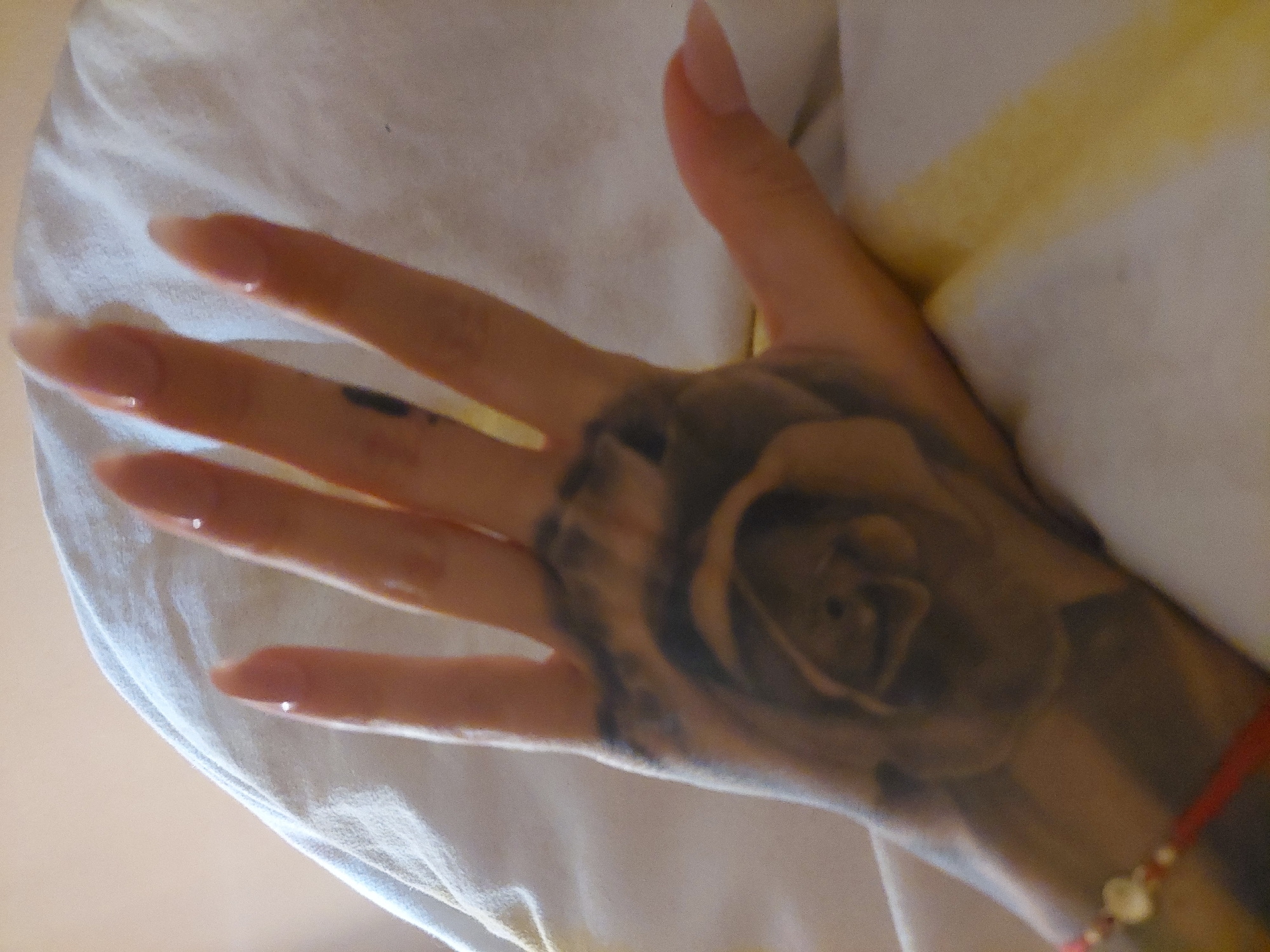 A close-up shot shows a woman's hand with a prominent rose tattoo spanning from the wrist to the back of the hand. A red string bracelet with small gold beads is visible around the wrist. The woman's fingernails are long and appear to be painted with a light pink or nude polish. A small, dark-colored mark is present on the finger between the index and middle fingers. The hand rests on a white and yellow striped cushion, likely indoors. There is no location in the city of Plovdiv, Bulgaria, visible in the image.