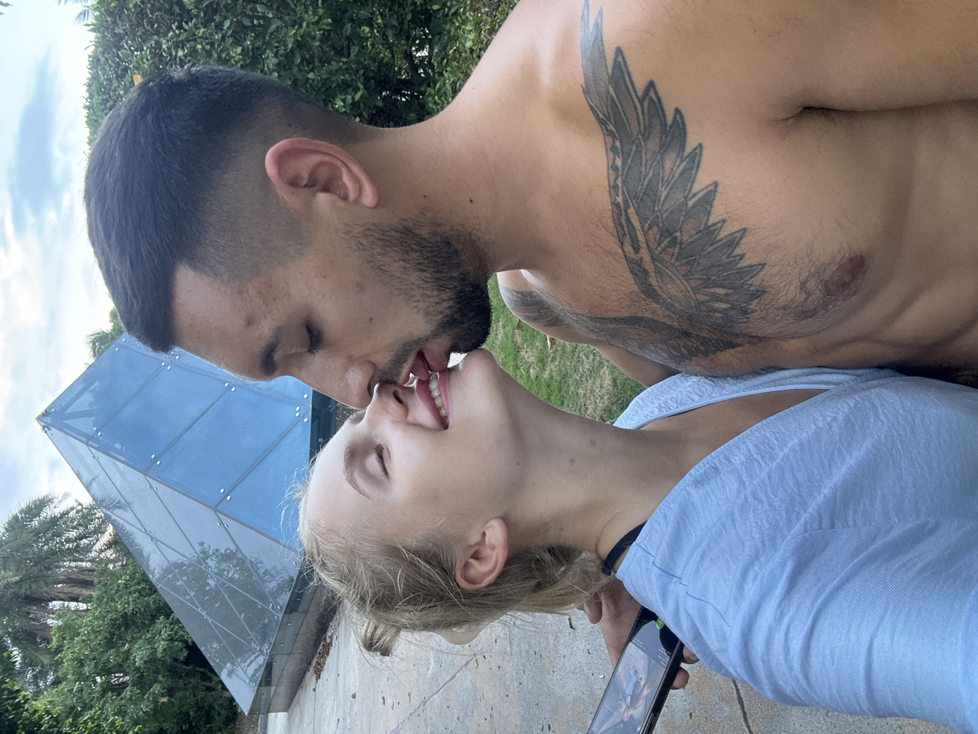 A couple is captured in a close-up embrace, with the man leaning in to kiss the woman's face. The woman has her eyes closed and her head tilted back slightly, a smile playing on her lips as his tongue touches her cheek. The man has his eyes closed as well, his beard visible. He has a large tattoo of wings on his chest. The woman is wearing a light blue top and has blonde hair tied back. She is holding a smartphone in her left hand, which is partially visible.
In the background, a modern, glass pyramid structure is visible, reflecting the cloudy sky. Lush green trees and foliage surround the area, suggesting an outdoor setting, possibly a park or a resort. The ground appears to be paved. This scene takes place in Nha Trang Ward, Vietnam.