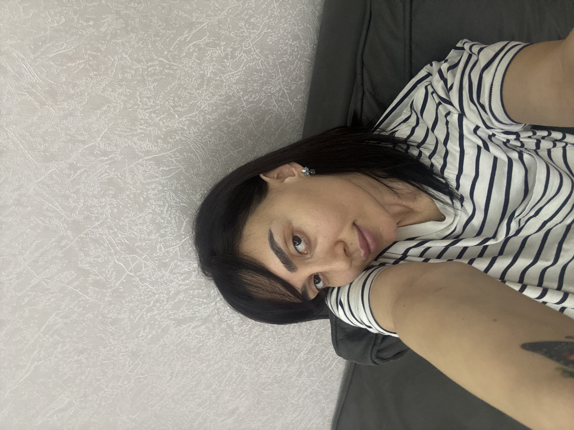 A woman is lying on a gray couch. She is wearing a striped shirt. The wall behind her is light gray. She is likely indoors. There is no information in the image to determine that the location is in the city of Tbilisi, Georgia.