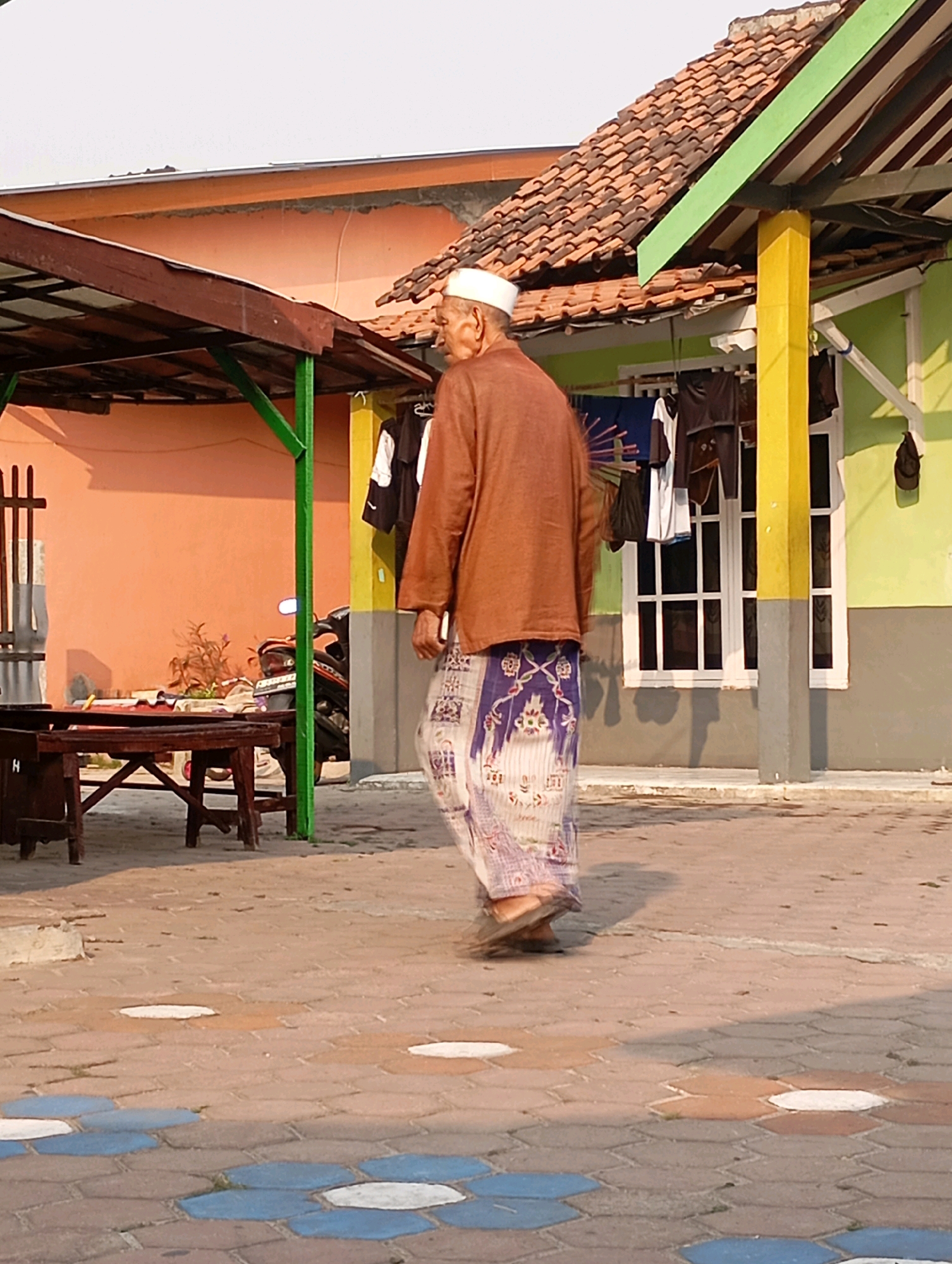 A man wearing a white cap, a brown long-sleeved shirt, and a patterned sarong walks on a paved courtyard in Babelan, Indonesia. He is facing away from the camera and to his right. Behind him, a light green building with a white window and a yellow support beam is visible. Laundry hangs on a clothesline in front of the building. To the left of the building, a pink wall with a wooden structure extending from it is present, featuring a wooden table and chairs. A motorcycle is parked behind the table. The courtyard is paved with hexagonal tiles in various shades of brown, blue, and white. The sky is overcast.