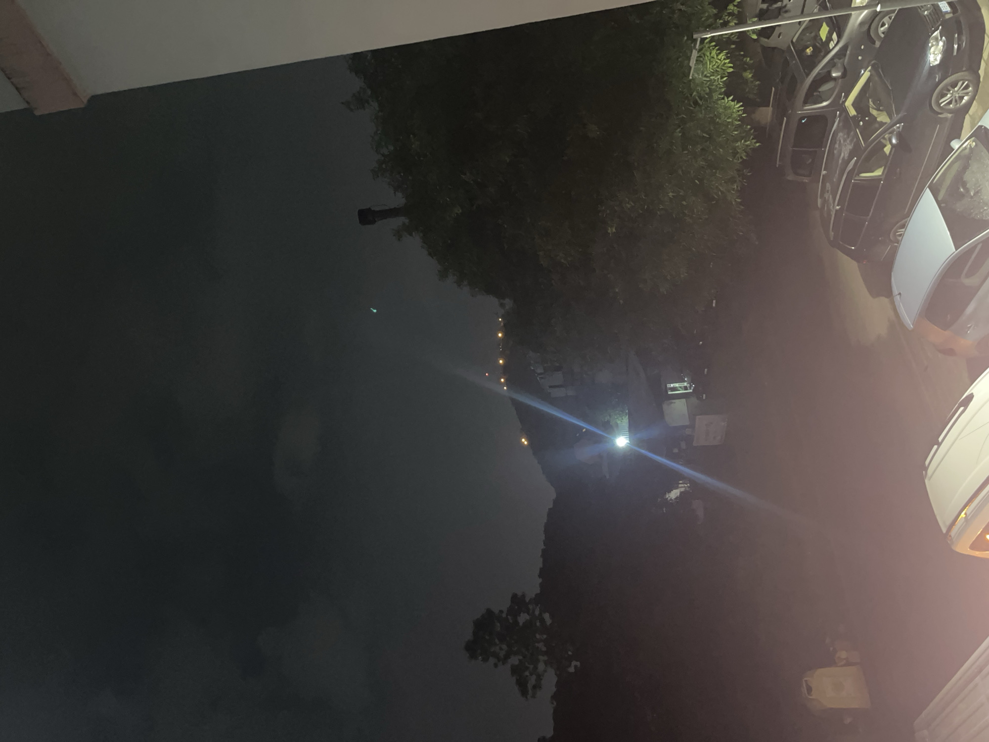 The image depicts a nighttime scene in Plovdiv, Bulgaria, viewed from a slightly elevated perspective, likely a balcony or window. The foreground is dominated by the edge of a building on the right, and the hoods of parked cars, including a light blue hatchback and a dark-colored station wagon. In the middle ground, a dimly lit area appears to be a parking lot or courtyard, with a few visible objects such as a trash receptacle and possibly some construction materials. Trees and foliage are prominent, casting shadows and obscuring parts of the scene. A bright light source, possibly a streetlamp, casts a strong beam and a visible lens flare towards the center of the image. In the background, a hill rises, dotted with lights from houses or buildings, suggesting a residential area on the outskirts of the city. The sky is dark and cloudy, with no visible stars.