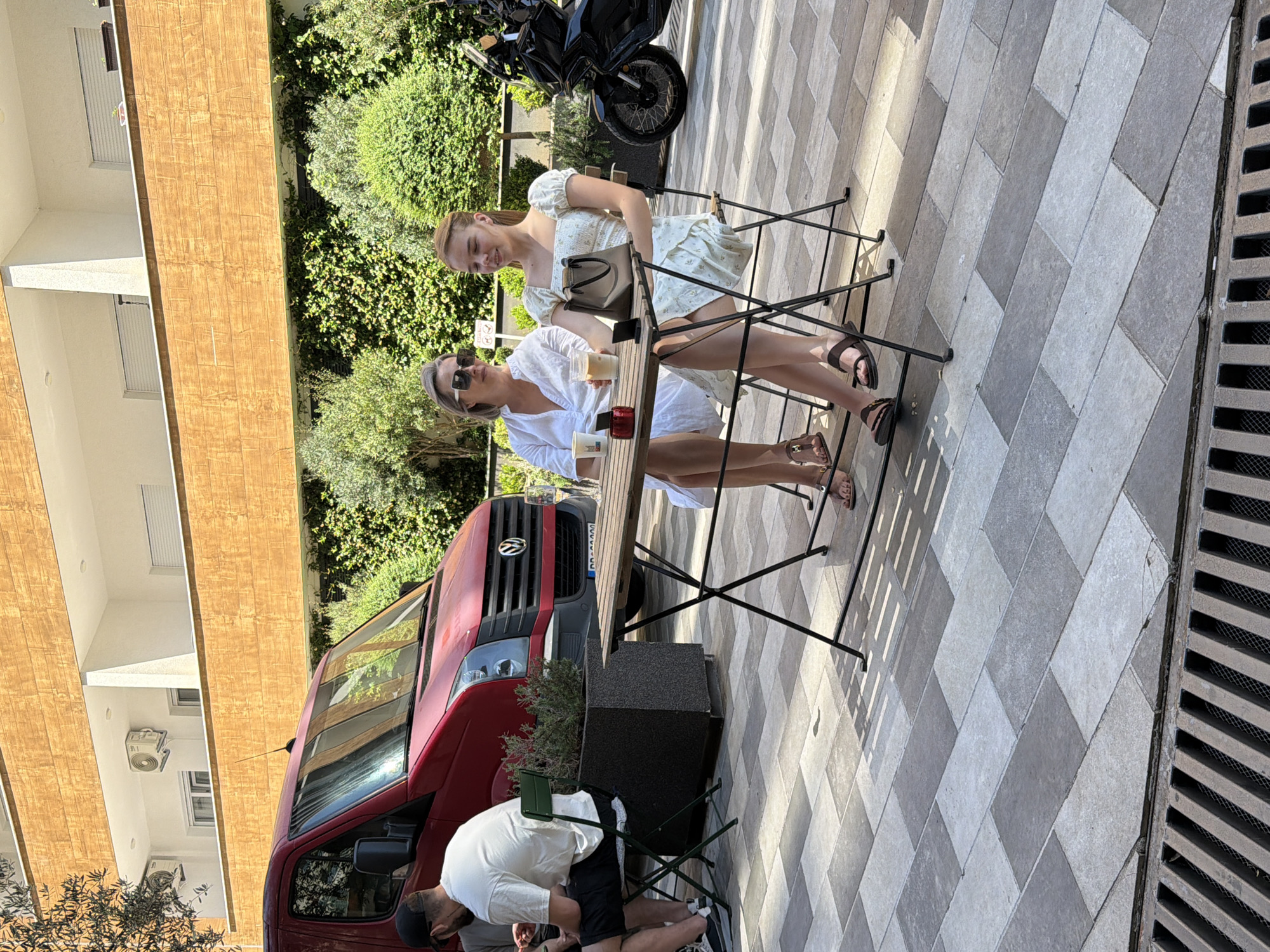 The image depicts an outdoor daytime scene in Budva, Montenegro, likely on a paved patio adjacent to a building. In the mid-ground, two women are seated at a square black metal folding table with a wooden top, facing each other in conversation. The woman on the left, appearing middle-aged, wears a white V-neck blouse, large dark sunglasses, and light brown sandals; she holds a white cup. The younger woman on the right wears a light-colored, short-sleeved dress and dark sandals, holding a clear glass containing a beverage. A light brown handbag and a small red object are placed on their table.
To the left, partially obscured, a man wearing a white t-shirt, dark shorts, and a black baseball cap is seated on a green folding chair at another table. Behind him, a red Volkswagen Crafter van is parked, visible from the front-right quarter, displaying a Montenegro license plate with "DD" visible. Further to the right in the background, a black motorcycle is partially visible.
The ground is paved with rectangular light gray and dark gray tiles arranged in a diagonal pattern. A metal drainage grate is present in the immediate foreground. The background features a multi-story building with white walls and balconies, characterized by horizontal wooden cladding. Air conditioning units are mounted on the balconies. Lush green foliage, including various shrubs and trees, borders the area behind the vehicles and tables. The scene is brightly illuminated by direct sunlight, casting distinct shadows.