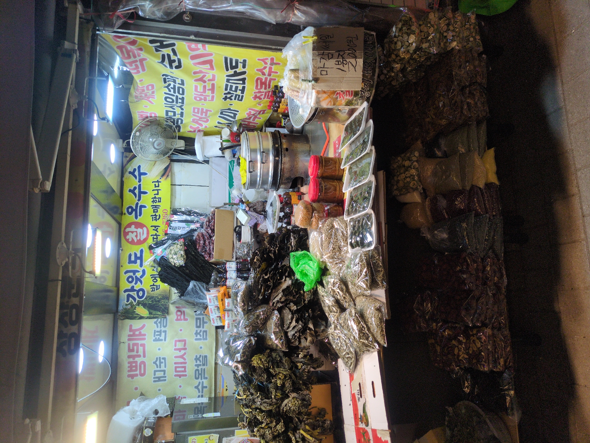 The image shows a street stall in Seoul, South Korea, selling various dried goods and prepared foods. There are multiple signs with Korean text advertising the products, including "Gangwon-do corn" and "Wando seaweed". The stall is stocked with dried vegetables, herbs, and seaweed, all neatly arranged. Several containers hold prepared foods, possibly kimchi or other fermented dishes. A small fan is visible near the top of the stall. The stall appears to be well-lit, and the environment suggests a bustling market or street food area.