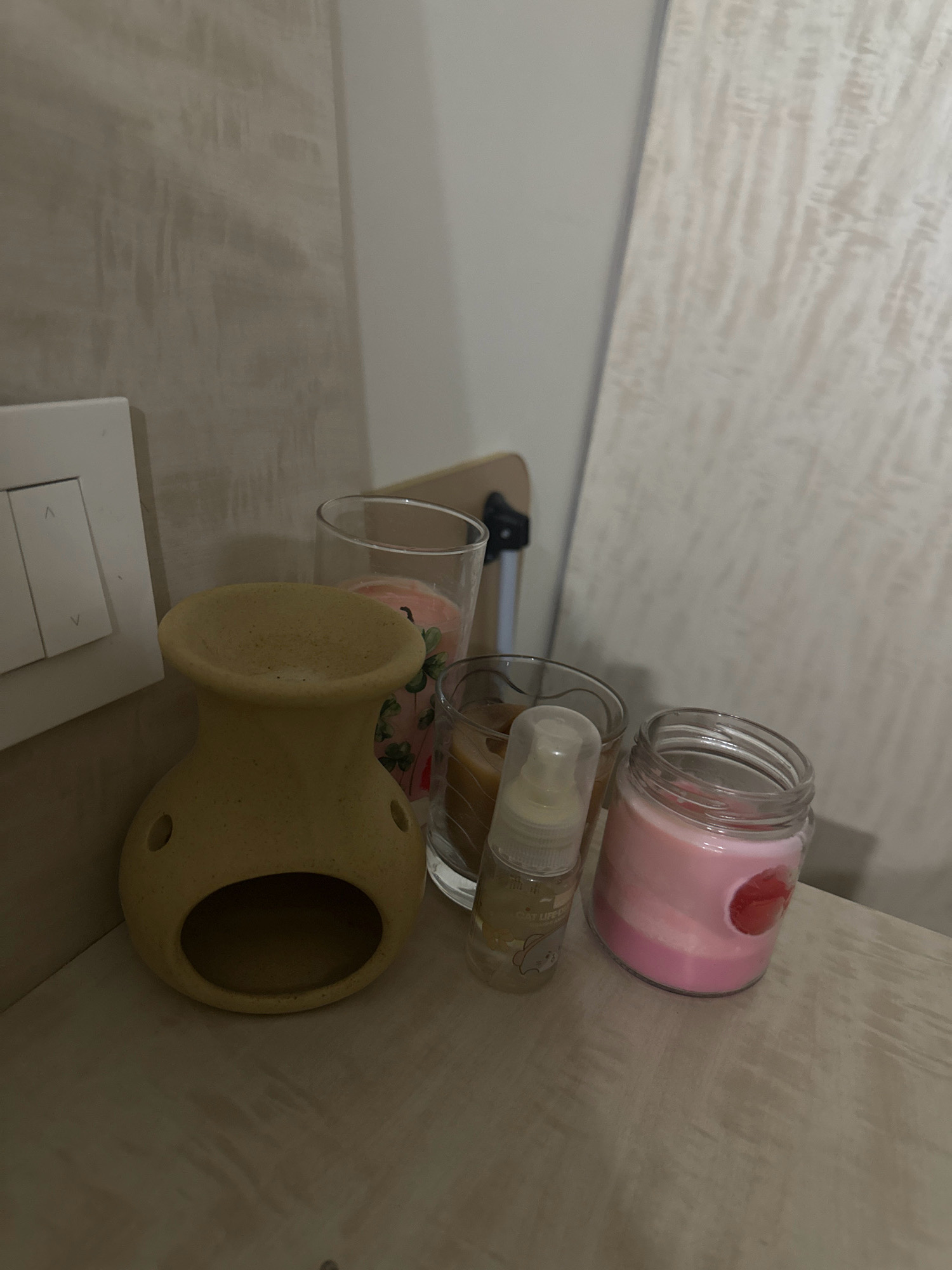A still life photograph taken at eye-level captures a collection of decorative items on a light-colored wooden shelf. The background consists of a textured wall with vertical lines and a white panel. On the shelf, from left to right, are a yellow ceramic oil burner, a clear glass with a candle inside featuring a floral design, a small clear spray bottle, and a pink candle in a glass jar. To the far left, a white light switch is visible on the wall. The lighting is dim, creating shadows and highlighting the textures of the objects. The scene is devoid of any people or animals. The photograph appears to be taken indoors, possibly in a residential setting. The location of Lalitpur, Nepal is not evident from the visual information provided in the image.