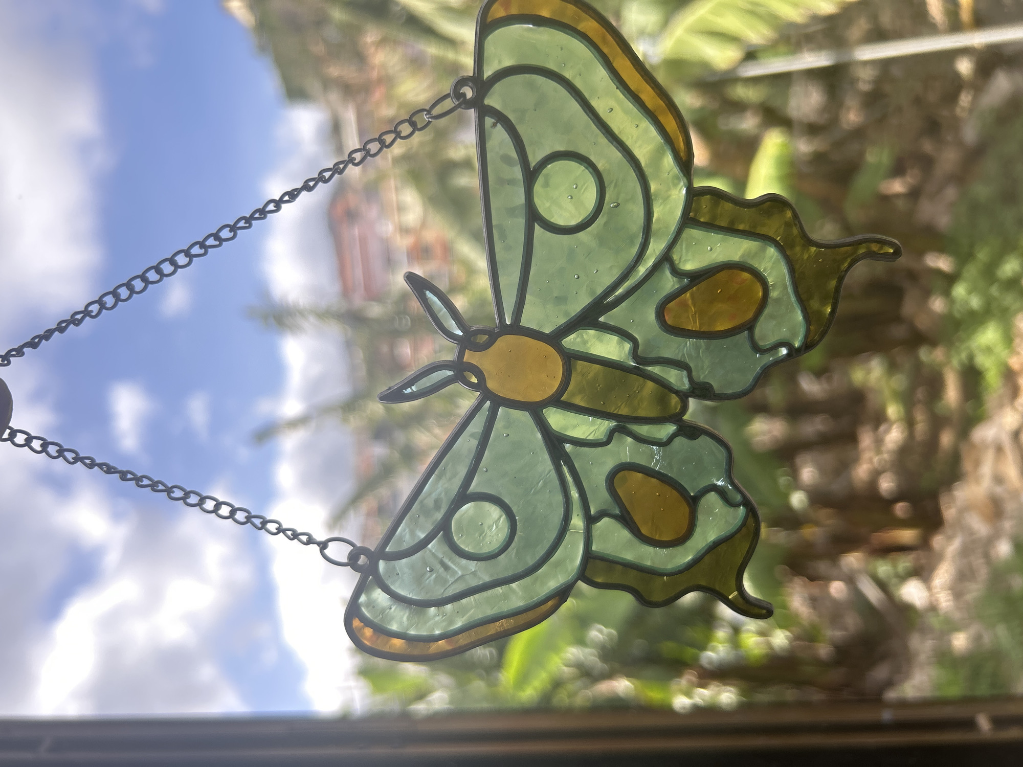 A close-up photograph showcases a stained-glass butterfly hanging in a window. The butterfly features green and yellow glass sections, outlined in black. It hangs by a metal chain. Outside the window, the blurred background suggests a daytime scene, with foliage and glimpses of buildings, possibly a residential area. The photograph does not contain information on people, actions, or interactions and the location is impossible to define from the image.