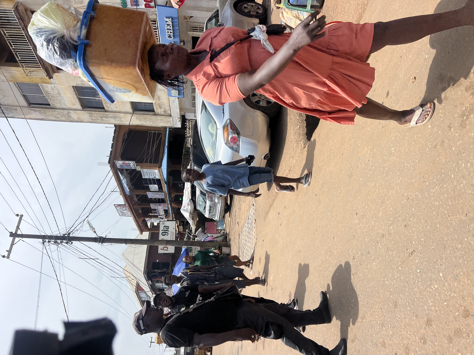 This media file shows a street scene in Nkpor, Nigeria. A woman in an orange dress is walking, carrying a large brown container filled with items on her head. She has a bag slung over her shoulder and is wearing white flip-flops. The scene appears to be during the day, under bright sunlight, casting long shadows on the dusty, paved road.
Several other people are also visible on the street, walking in various directions. A young boy in a blue shirt and shorts is walking ahead of the woman, and further down the road, a group of men in dark clothing are walking together. Some appear to be looking towards the camera. In the background, there are buildings, a white car, and a utility pole with many tangled wires. The buildings seem to be shops, with some signage visible. One sign reads "9ja". The overall atmosphere suggests a busy commercial or residential street.