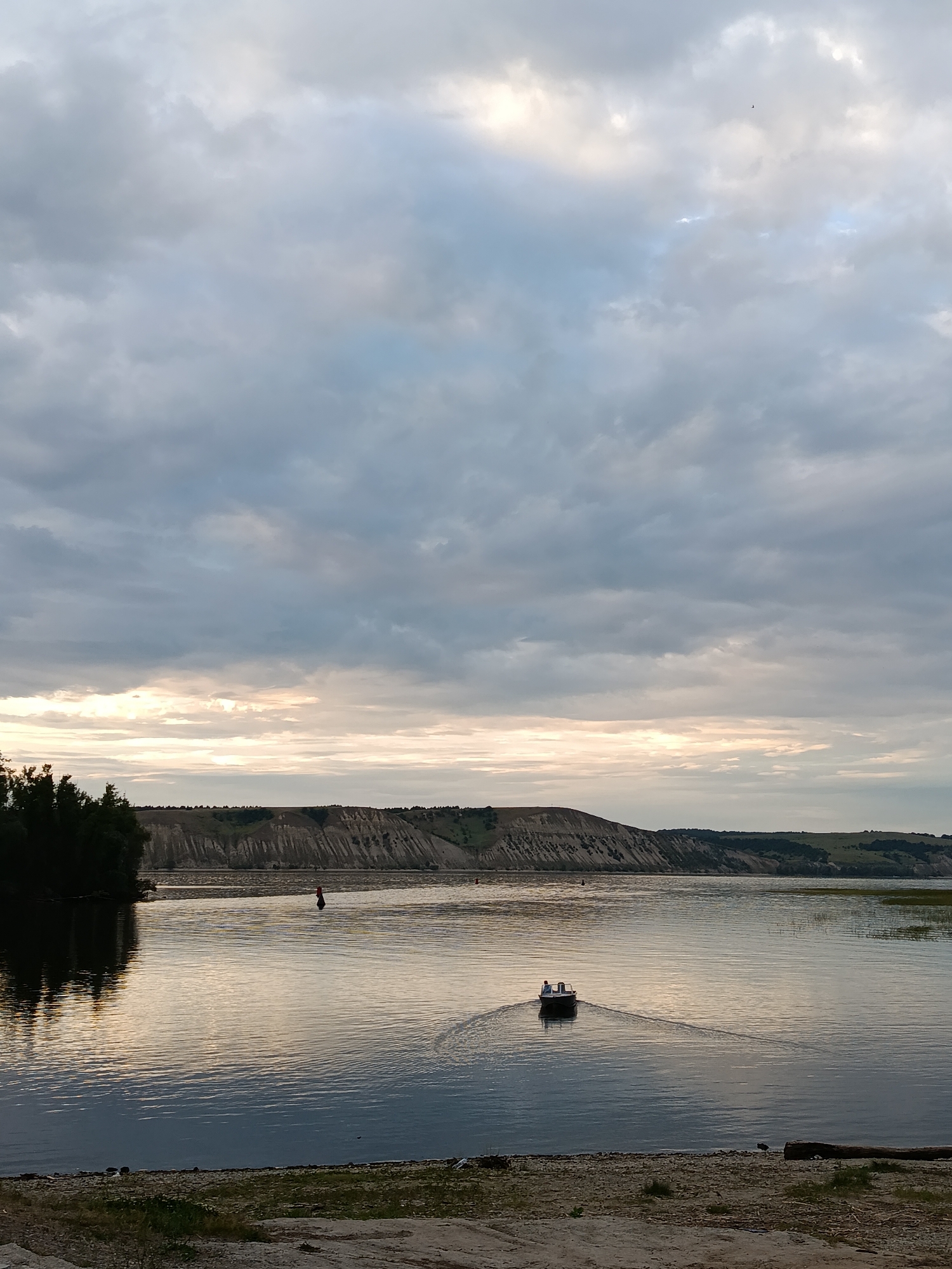 The image shows a wide river with a small boat containing two people in the middle distance. The boat is moving away from the viewer, leaving a wake on the water. On the left bank of the river, there are dark green trees with their reflections visible in the water. Across the river, there is a gently sloping hill with a textured, uneven surface, possibly a cliff face or eroded landform, dotted with vegetation. The sky is overcast with a mix of grey and lighter clouds, with a band of pale yellow light visible on the horizon, suggesting the sun may be setting or rising. The foreground consists of a sandy bank with some sparse vegetation and a fallen log. The overall scene suggests a natural landscape, likely in a rural or semi-rural area. This image was taken near the city of Marx, Russia, on the Volga River.