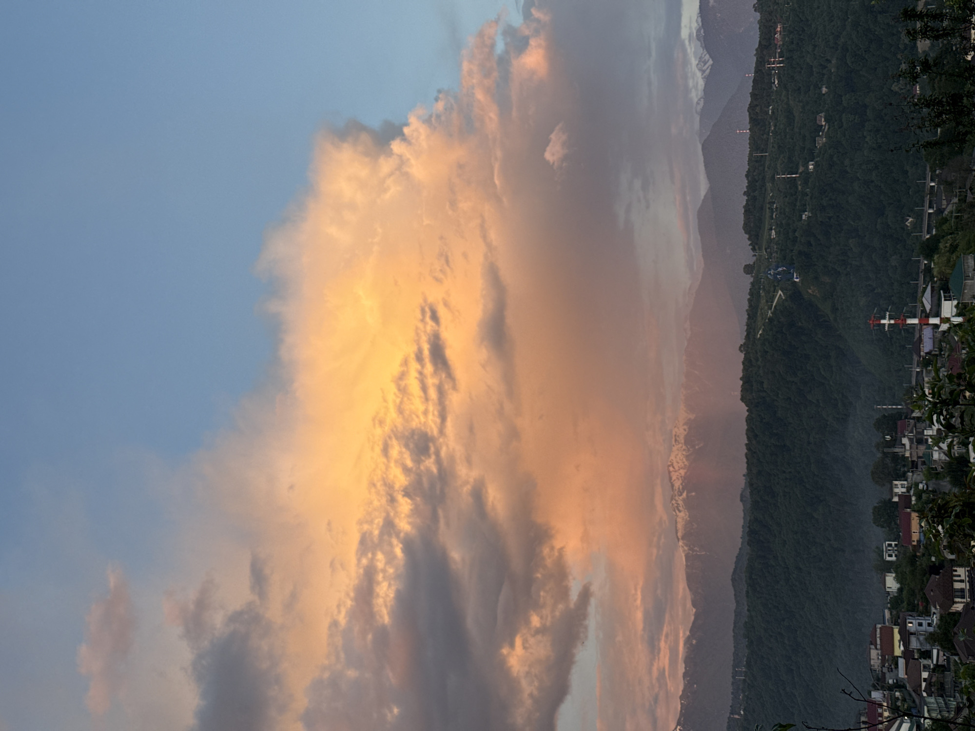 The image shows a view in Sochi, Russia, featuring a large, illuminated cloud formation dominating the left side of the frame. The sky transitions from blue to orange-pink hues due to the setting sun. In the background, rolling green mountains and snowy peaks are visible. The right side of the image shows a hillside dotted with buildings, trees, and what appear to be antennas or towers.