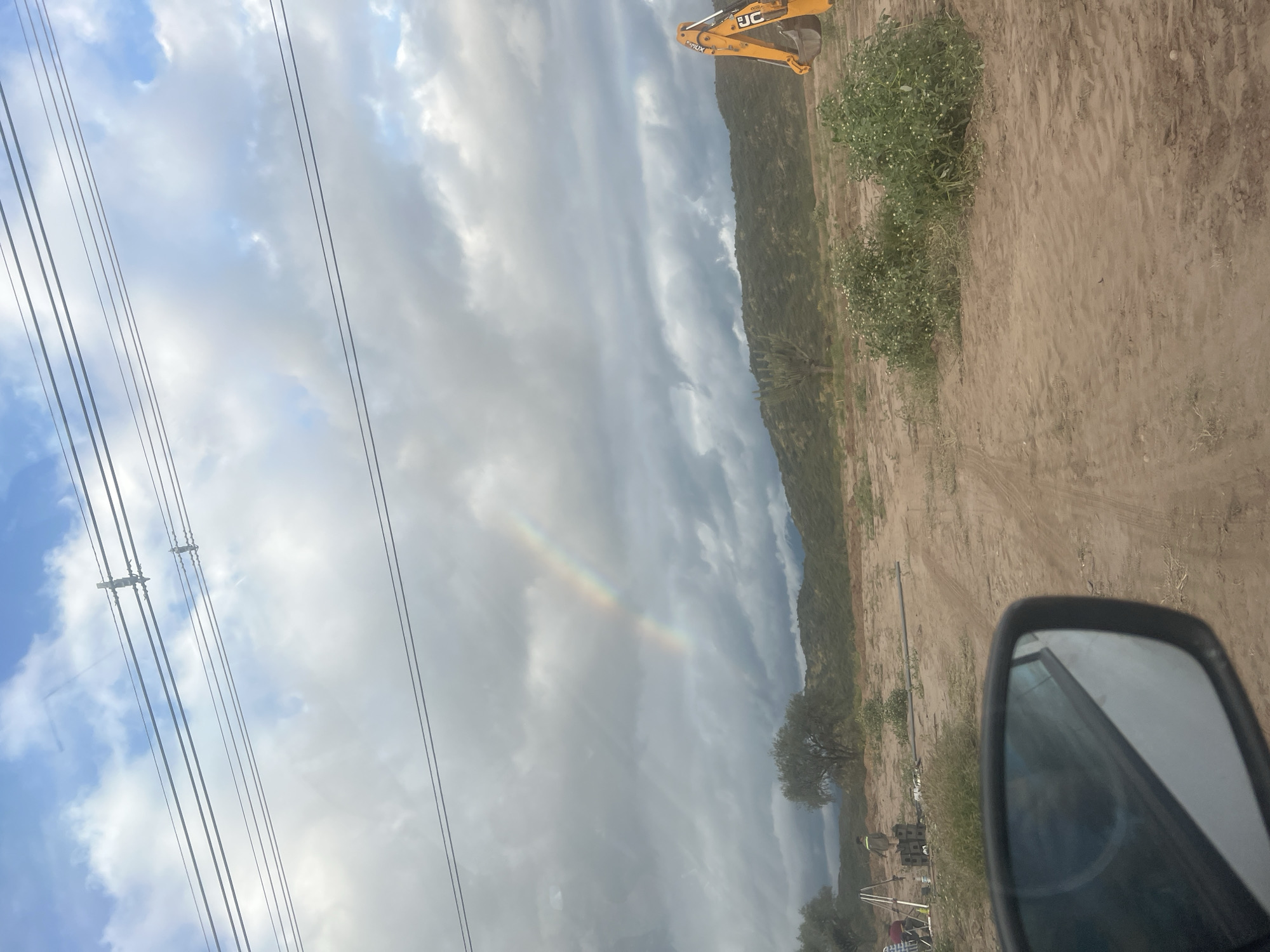 The image shows a landscape with a combination of natural and human-made elements. In the sky, there are power lines extending across a partly cloudy sky with a faint rainbow visible. The terrain appears to be arid, with a mix of dirt, sparse vegetation, and what looks like a hillside or low mountain range in the background. A piece of yellow heavy machinery is positioned on the terrain, likely used for construction or excavation. A side mirror of a vehicle is visible, suggesting the photo was taken from inside a car. There are no people definitively identifiable. This image may be in San Fernando del Valle de Catamarca, Argentina, but this cannot be confirmed without further information.