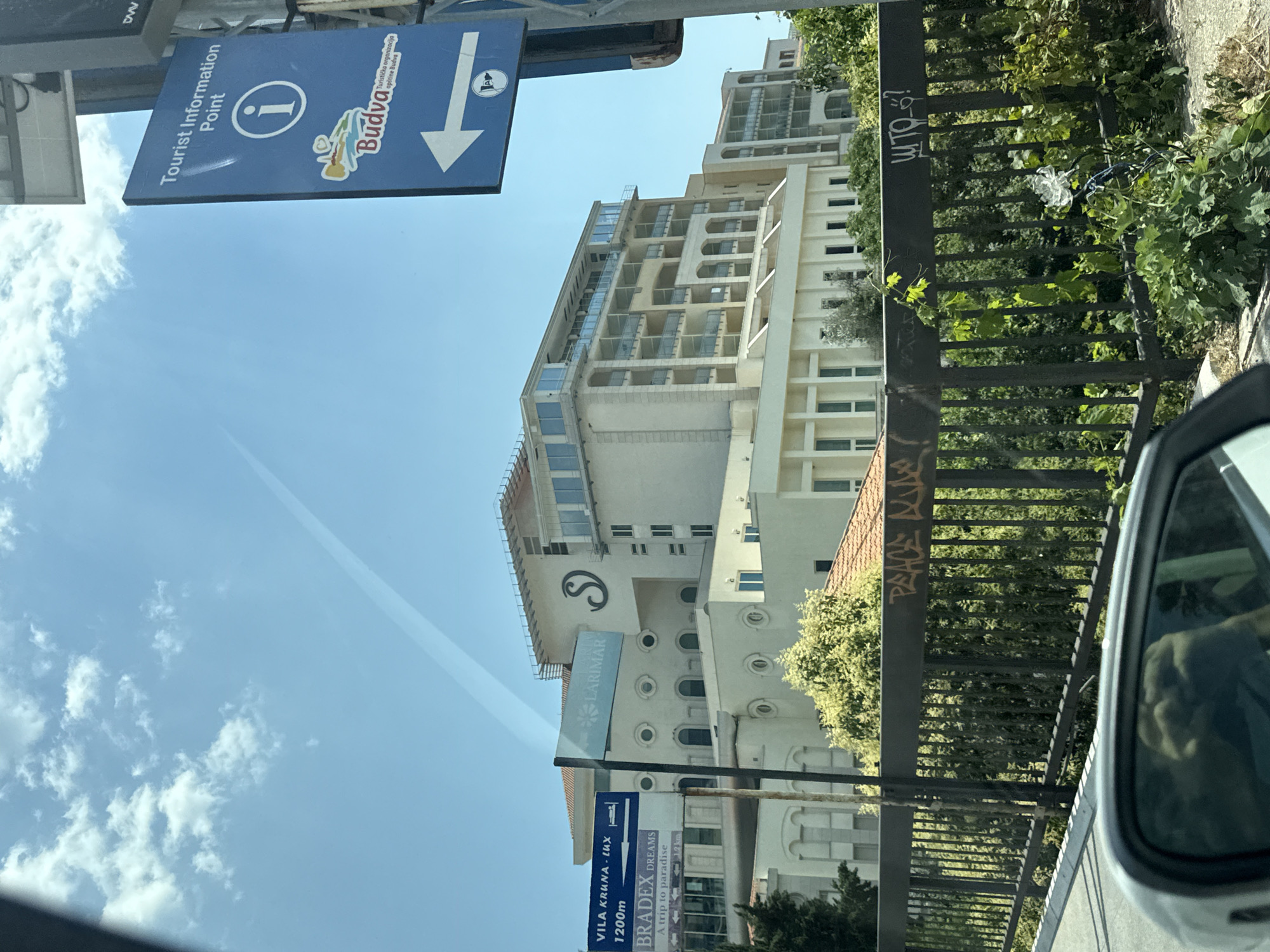 The image depicts a sunny day in Boreti, Montenegro. In the foreground, a car's side mirror reflects a person's face. A black metal railing runs across the middle ground, adorned with graffiti. Behind the railing, lush green trees and foliage are visible, with a large building complex dominating the background. The building is white with multiple floors, featuring balconies and numerous windows. Several signs are present: one blue sign on the right indicates "Tourist Information Point" with an informational icon and a logo for "Budva." Another blue sign on the left displays directions to "VILA KRUHA - LUX 1200m" and "BRADEX DREAMS A trip to paradise." A third sign, partly obscured, reads "LARIMAR." The sky is a clear blue with scattered white clouds.